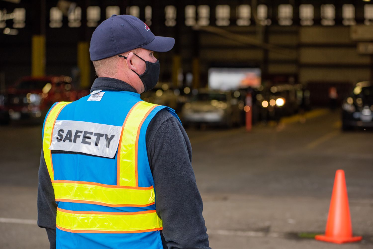 A masked worker in a blue safety vest and cap stands in a vehicle processing area with an orange traffic cone nearby.