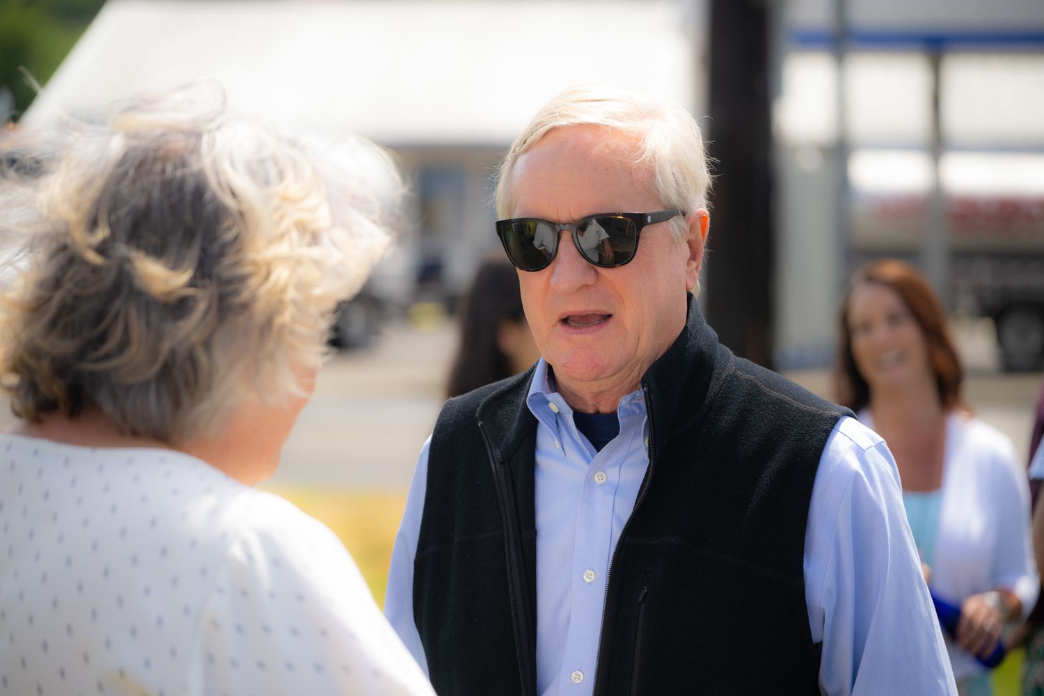 Older man in dark sunglasses, a black vest and light blue shirt talks with a woman at an outdoor event on a sunny day.