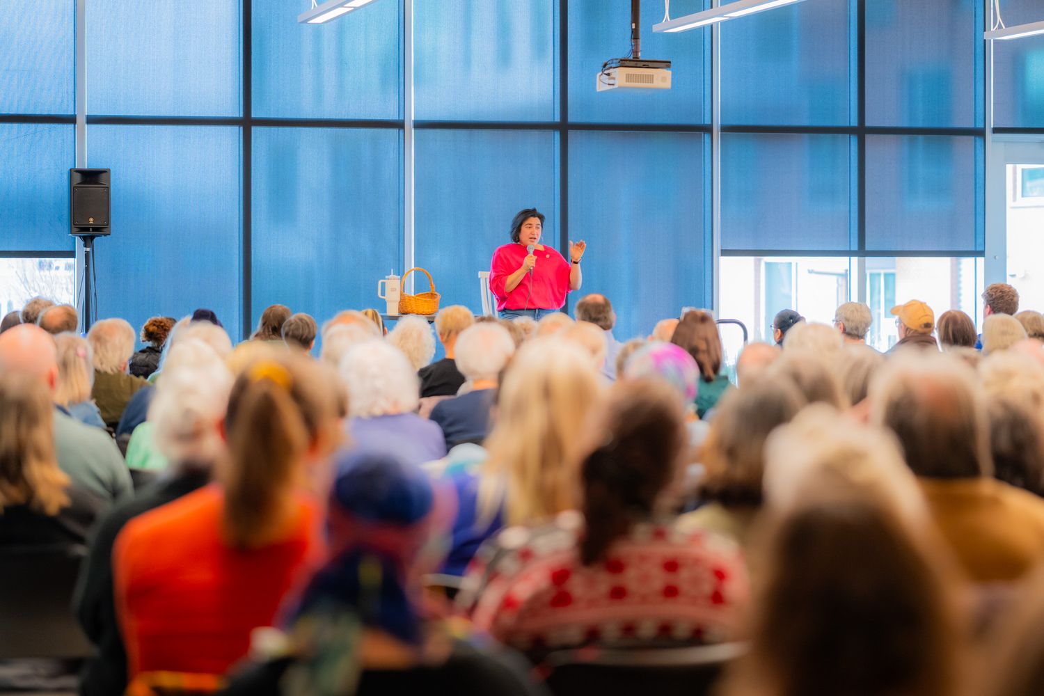 A woman in a bright red top speaks to a large seated audience in a room with tall windows, filmed from behind the attendees.