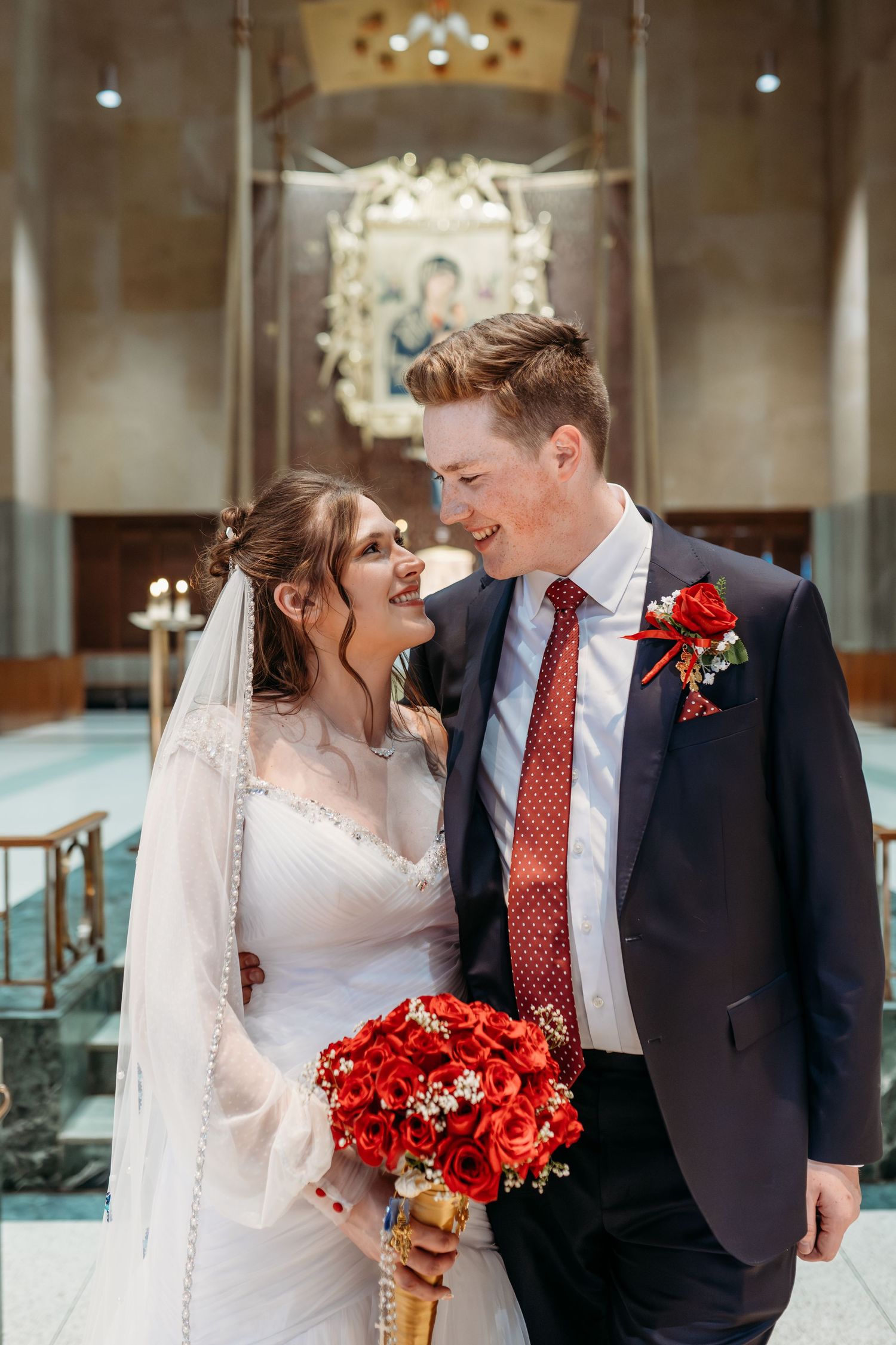 A bride and groom share a tender moment in a church interior with red roses and white decor.