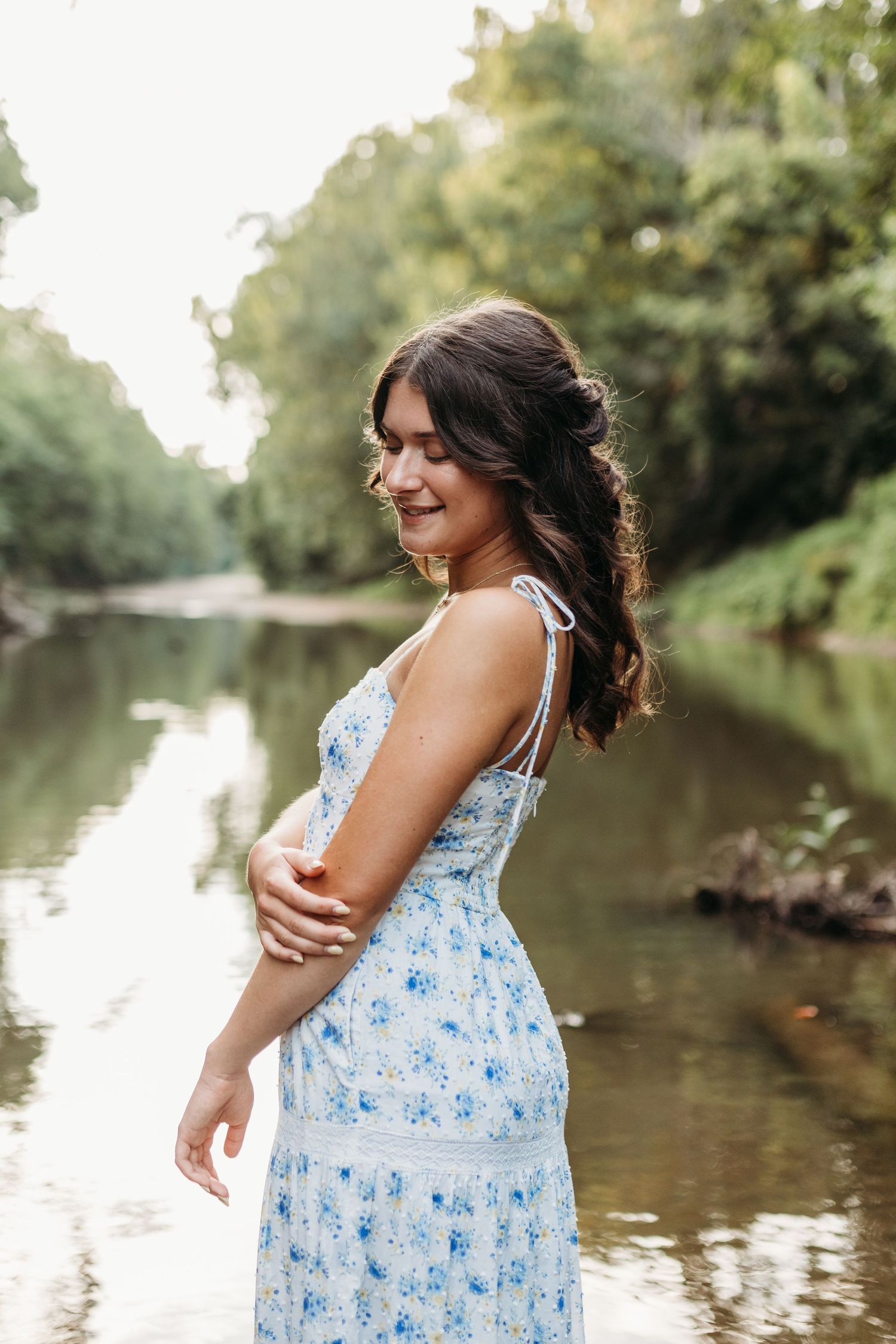 A senior getting her outdoor senior photos taken in St. Louis wearing a blue floral summer dress stands smiling by a river on a sunny day.