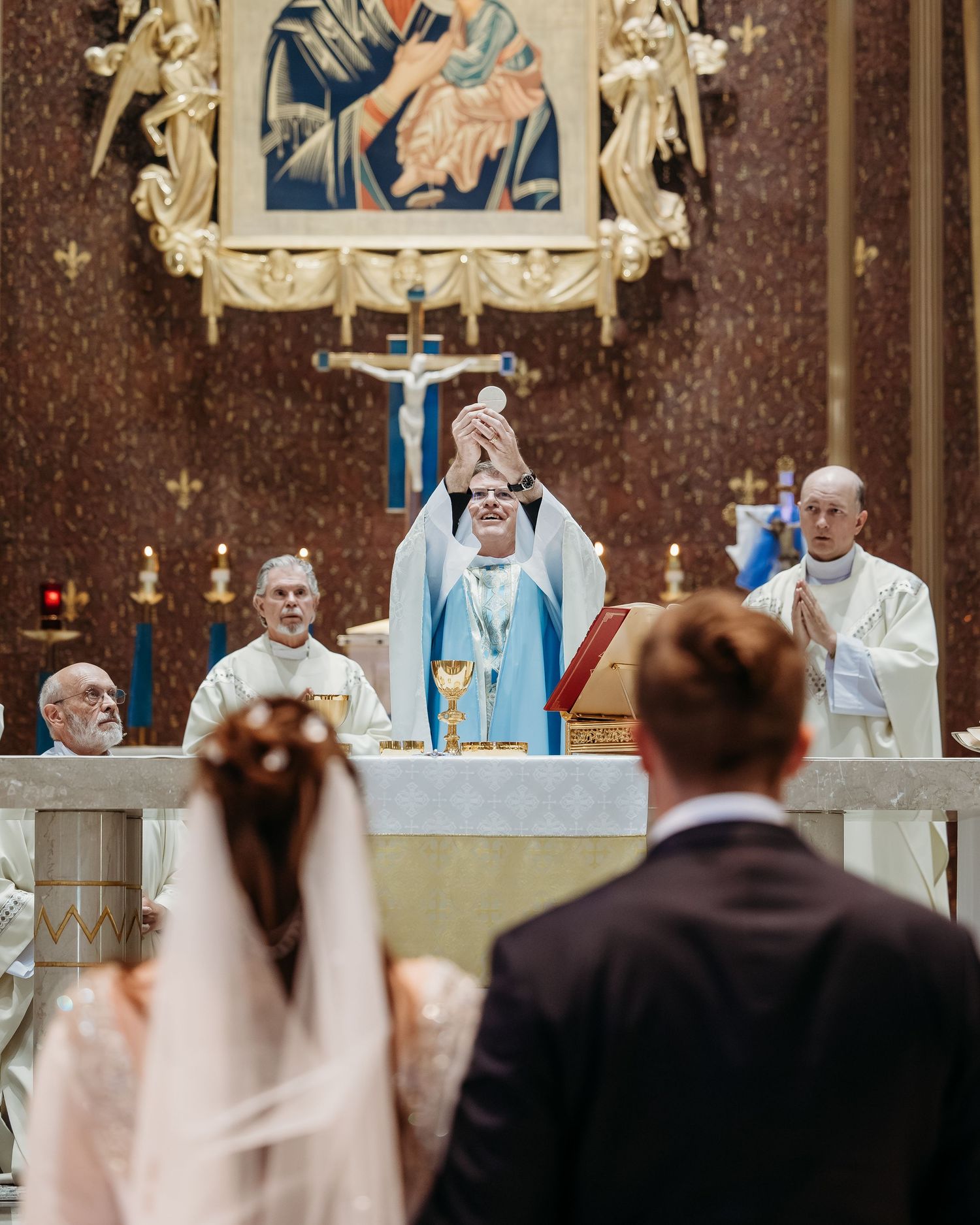 Clergy members perform a traditional Catholic wedding ceremony at an ornate church altar with religious artwork and candles.