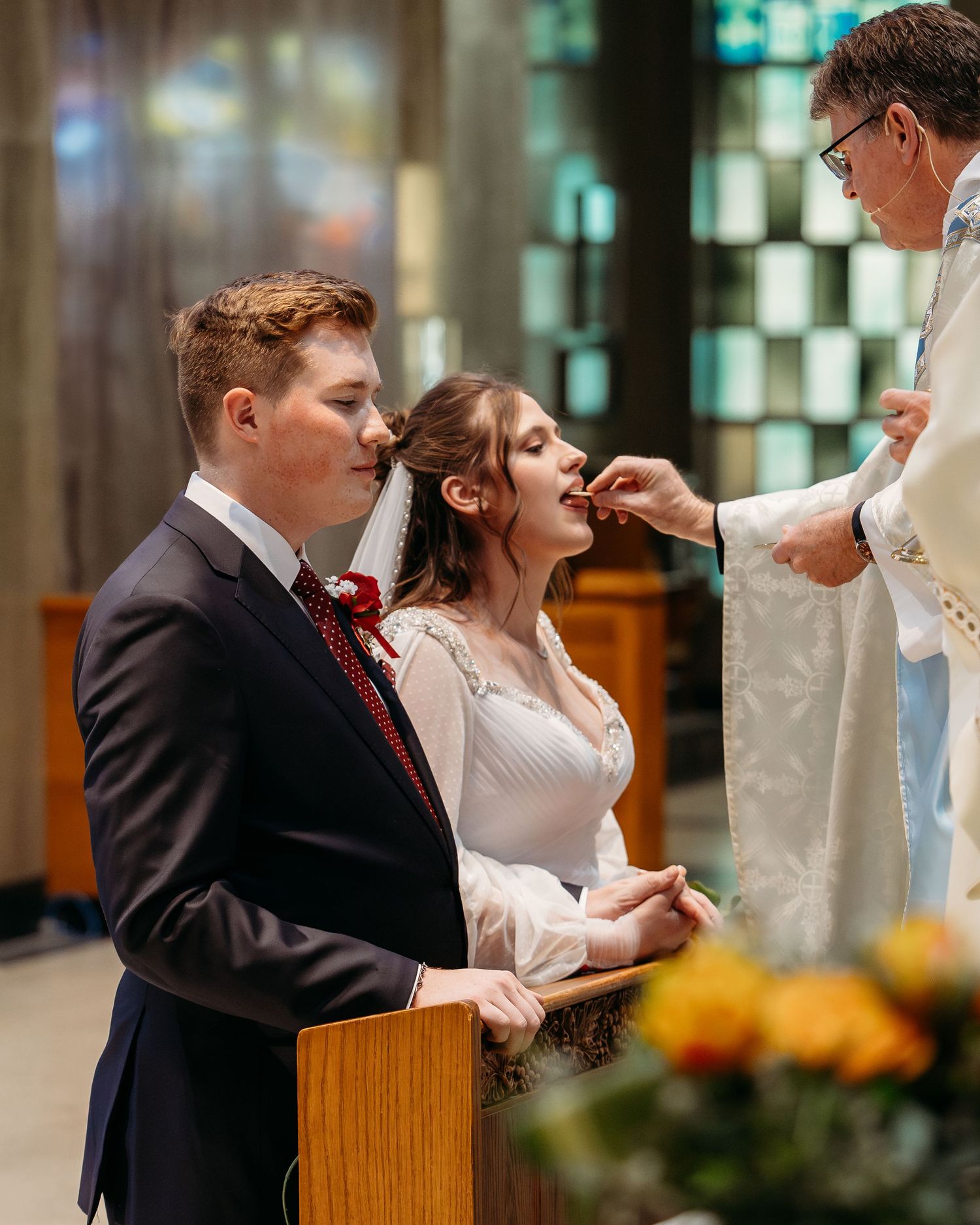 A couple participates in a traditional Catholic wedding ceremony as they receive communion from the priest at the altar.