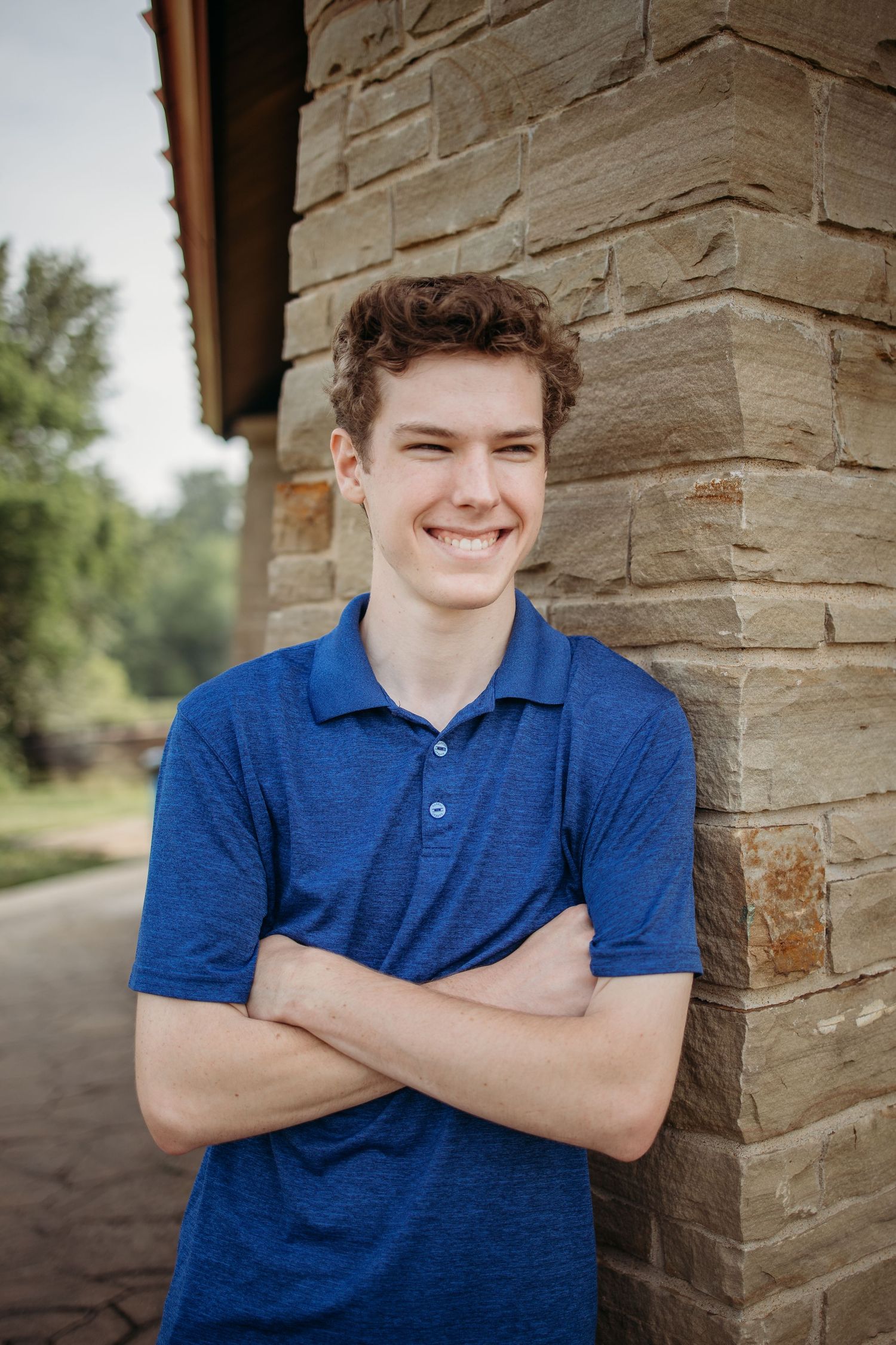 Senior wearing a blue polo shirt with natural light stands against a stone wall exterior, smiling warmly at the camera.