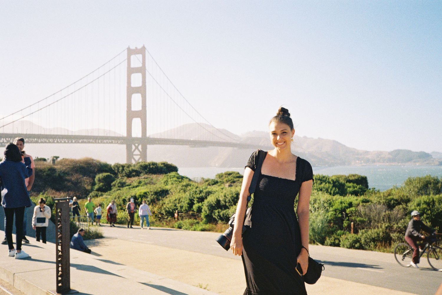 Woman in black dress holding camera with iconic Golden Gate Bridge in background on sunny day.