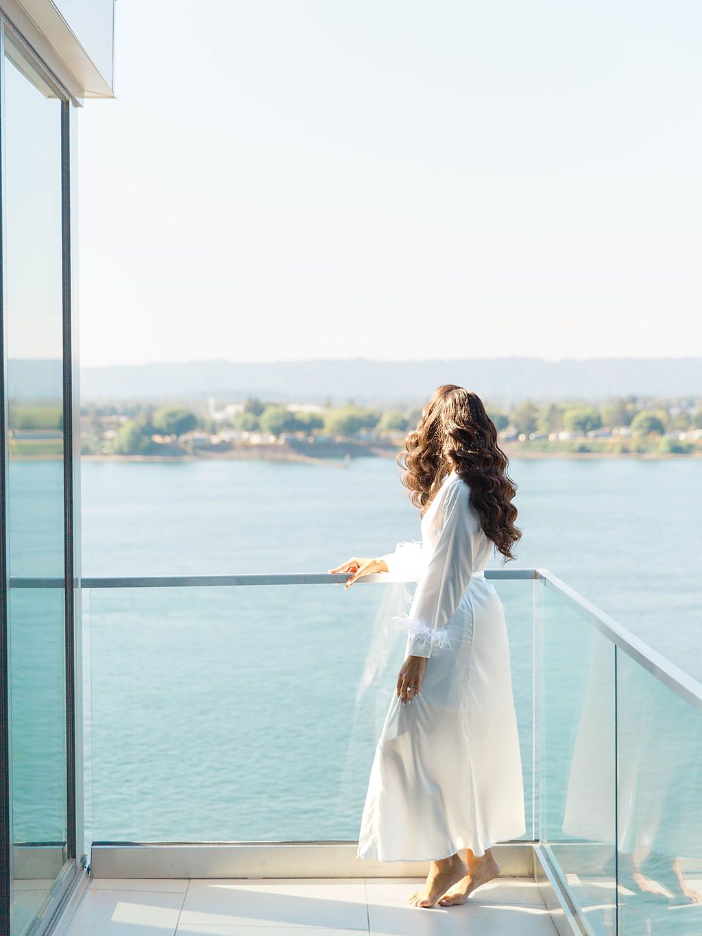 Bride with long curly hair in a white dress gazes at a scenic lake from a modern glass balcony.