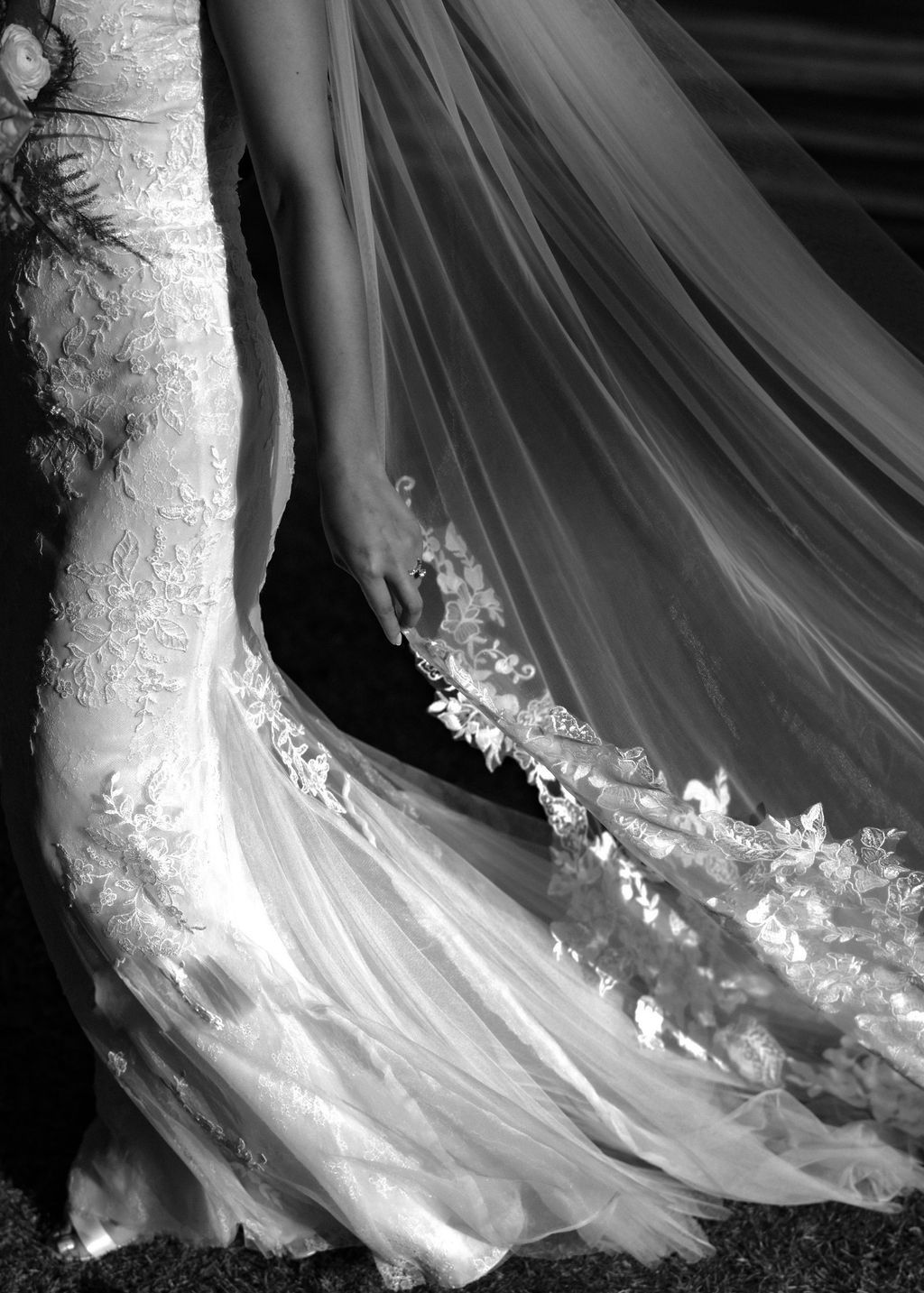 Bride in lace mermaid wedding gown with flowing floral-applique veil billowing in wind, black and white.