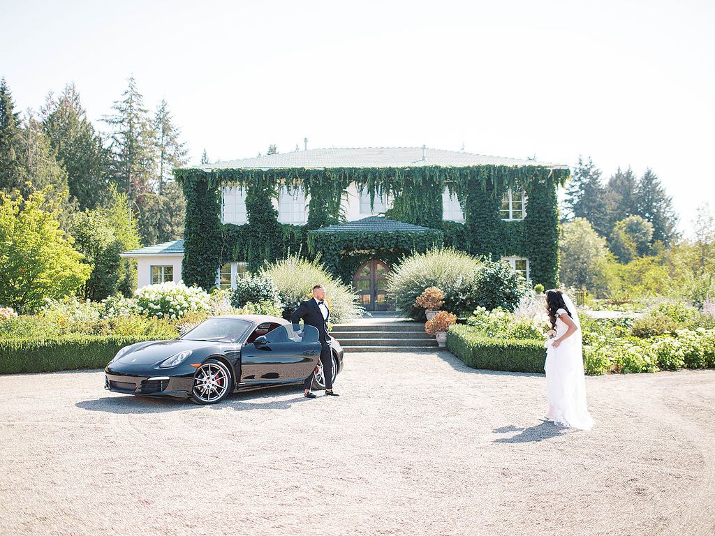 Bride in white gown and groom beside a black Porsche at an elegant ivy-covered estate venue.