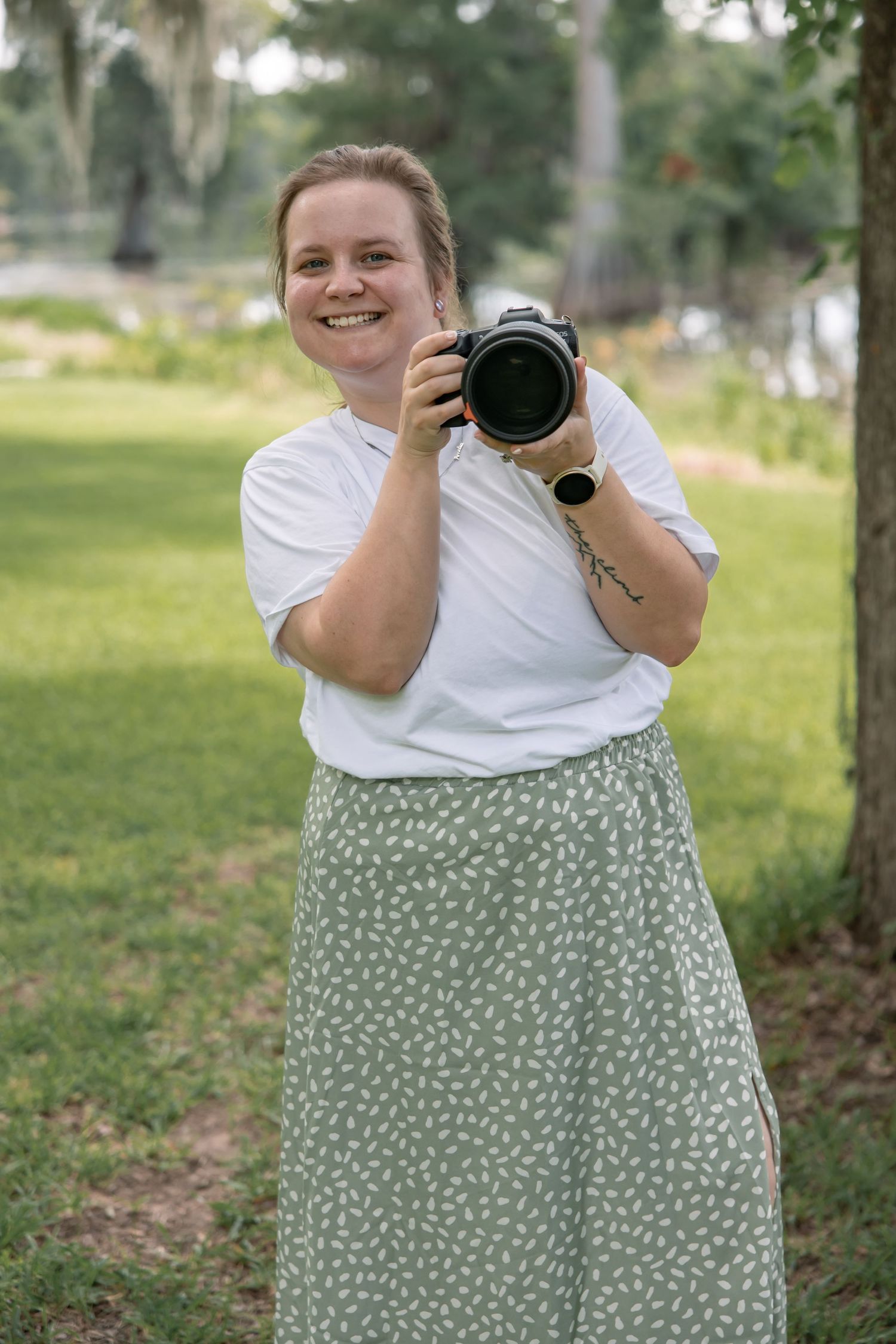 Smiling woman holding a DSLR camera outdoors in a green park, wearing a white tee and sage floral midi skirt.