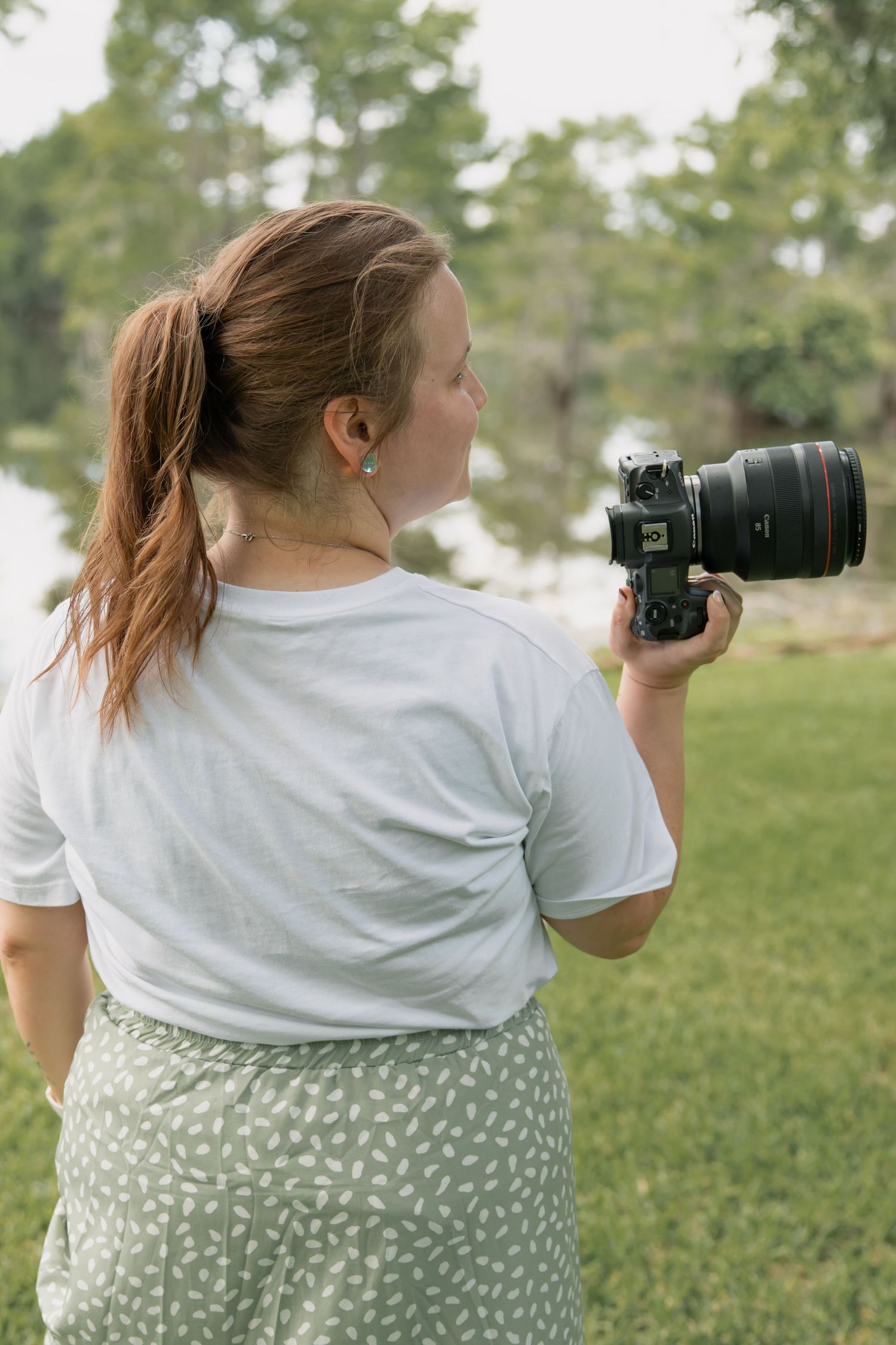 Woman in white t-shirt and green skirt holding a DSLR camera outdoors near a lake surrounded by trees.