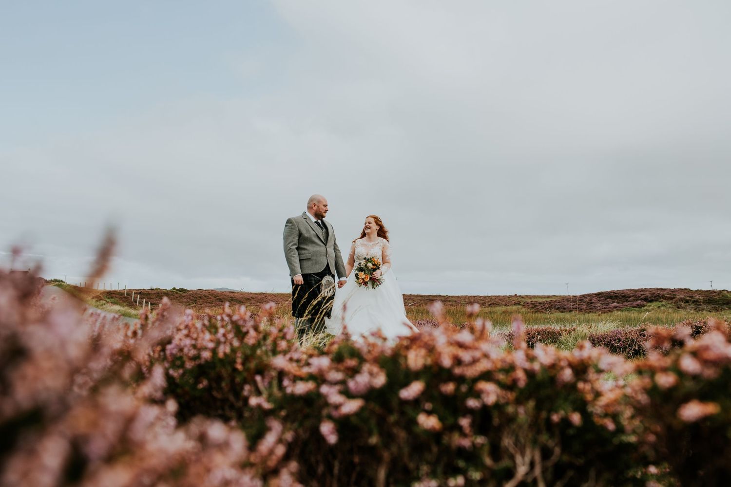 A wedding couple stands amid pink heather blooms on a misty hilltop.