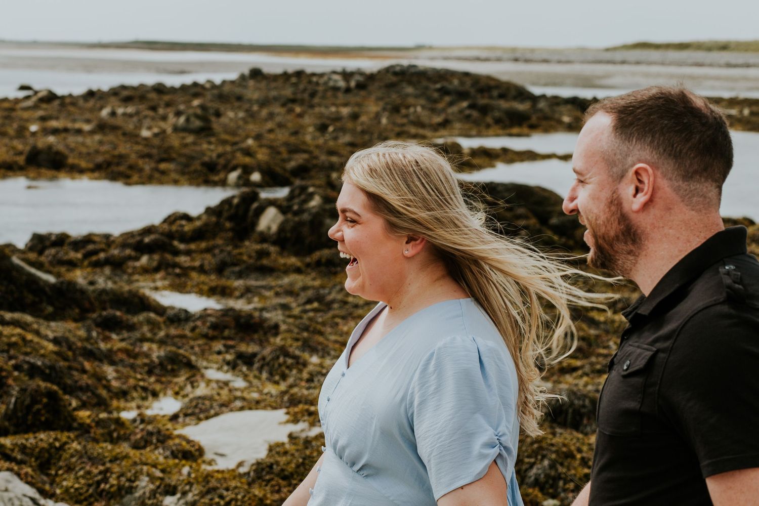 A couple laughs together on a rocky beach as wind blows through their hair.