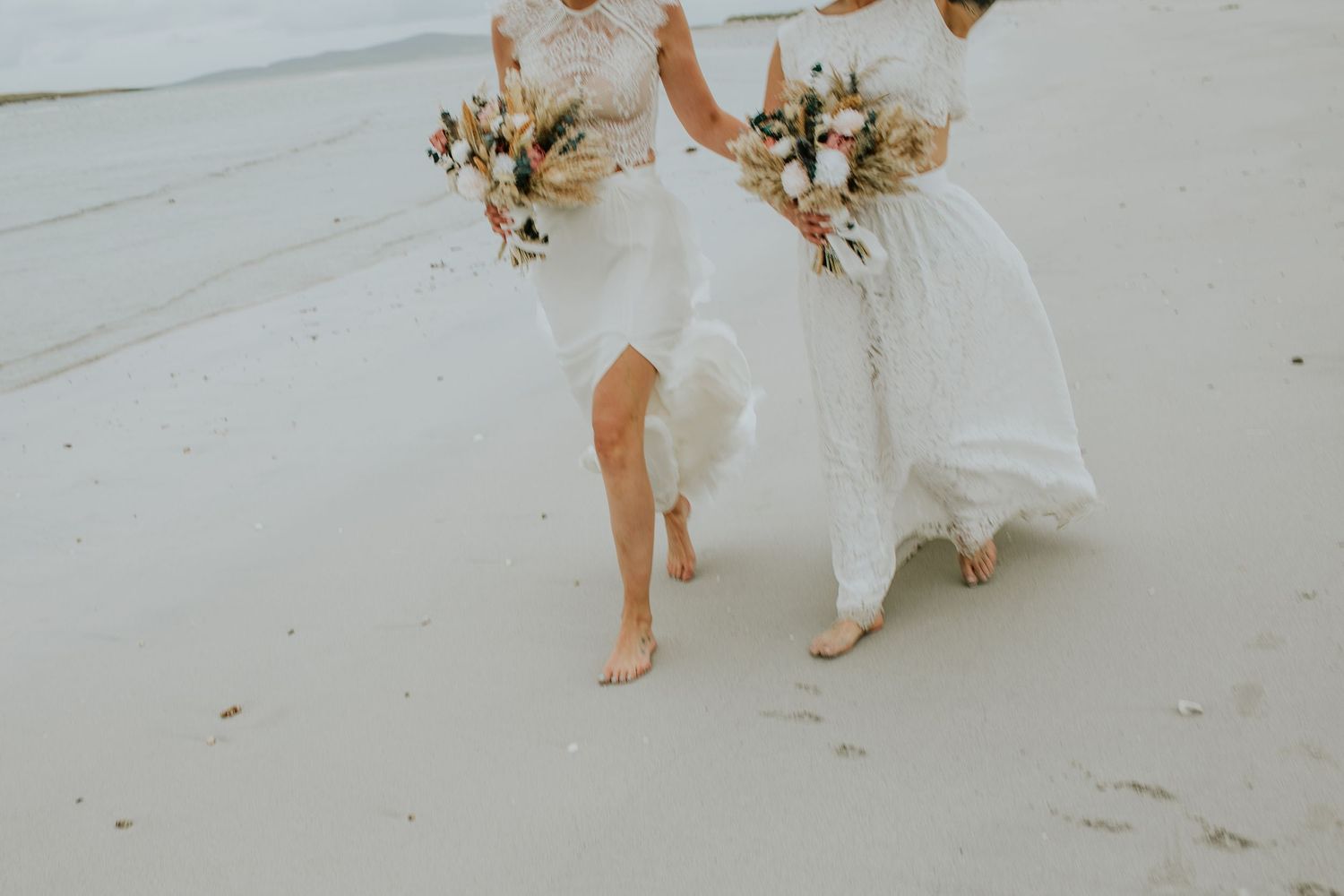Bare feet in white wedding dresses walk across a sandy beach holding neutral-toned bouquets.