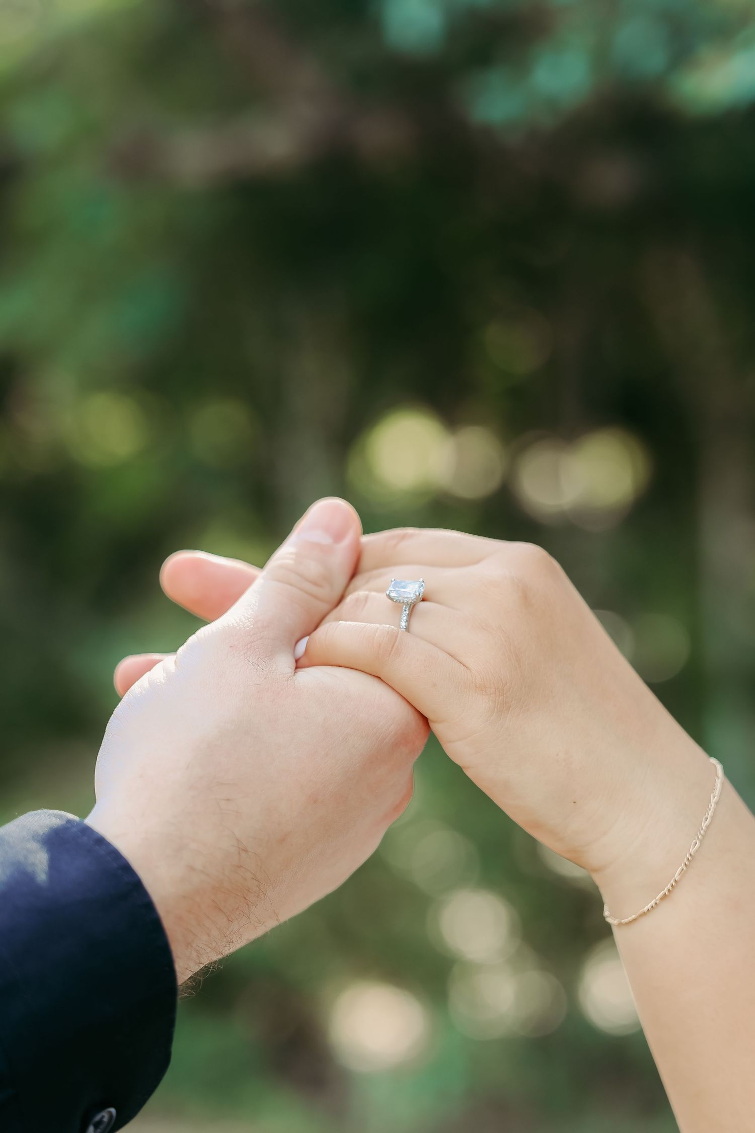 Close-up of hands holding with engagement ring in focus against blurred green background.