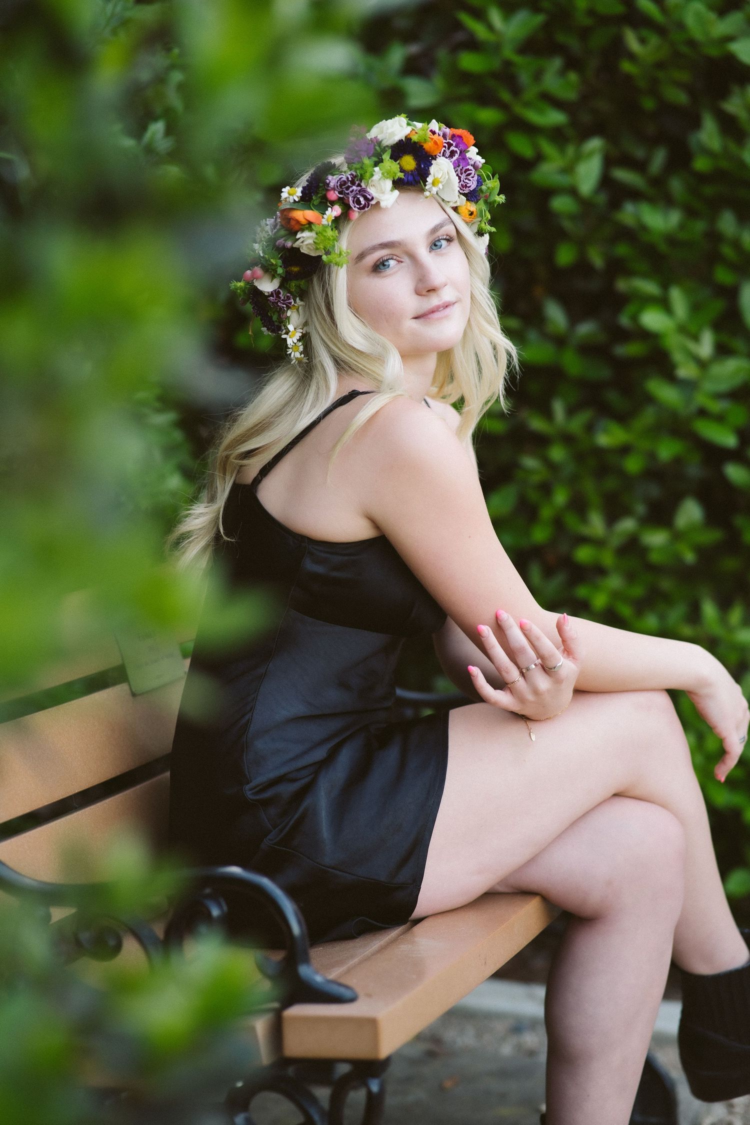 A dreamy outdoor portrait of someone wearing a colorful floral crown and black dress sitting on a bench.