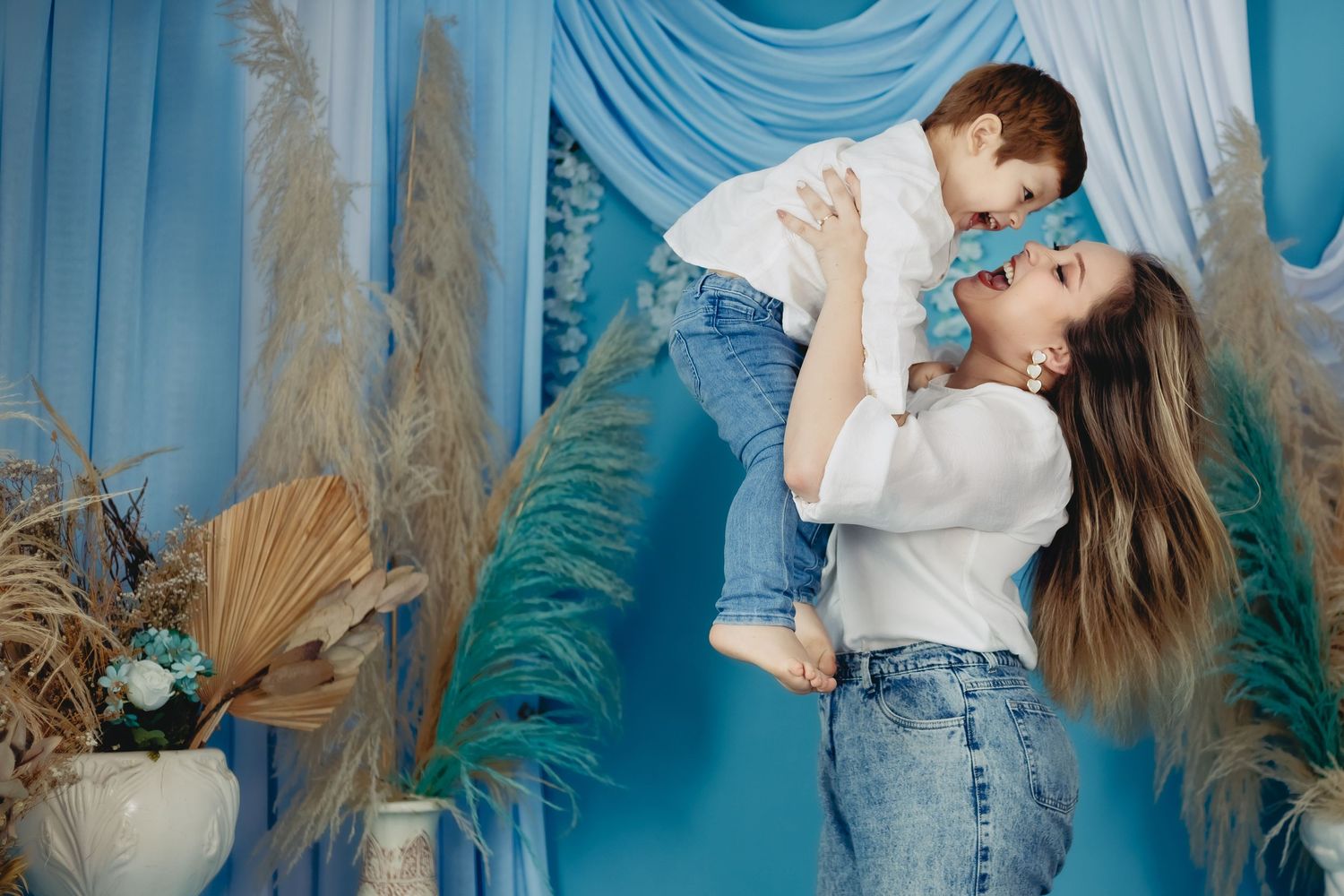 A mother joyfully lifts her young son in a blue-themed studio with dried pampas grass decor.