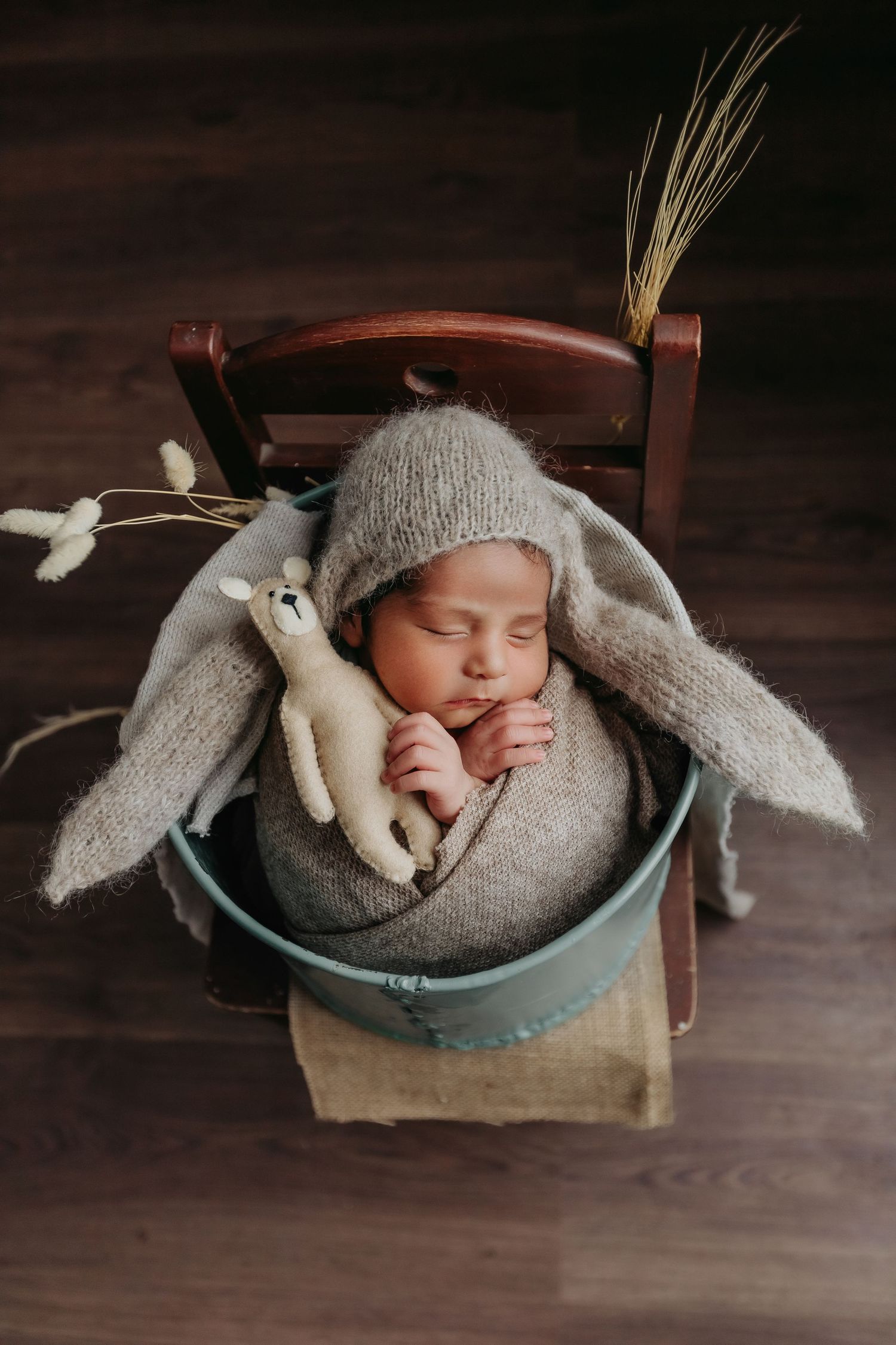 Sleeping newborn wrapped in gray knit blanket with bunny toy in vintage turquoise basket on wooden floor.