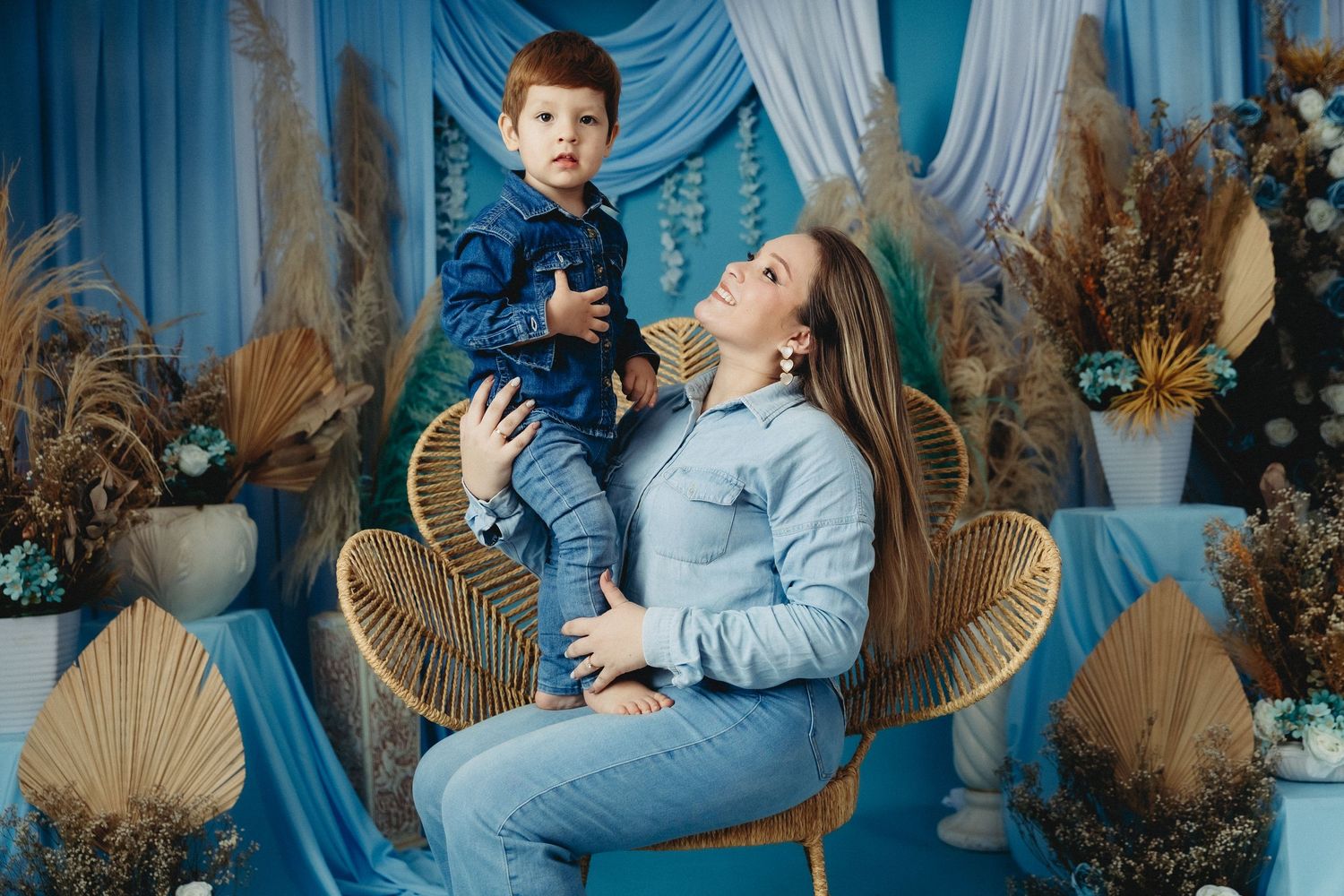 A woman sits in a wicker chair holding a toddler boy, both in denim, against a blue decorative backdrop.