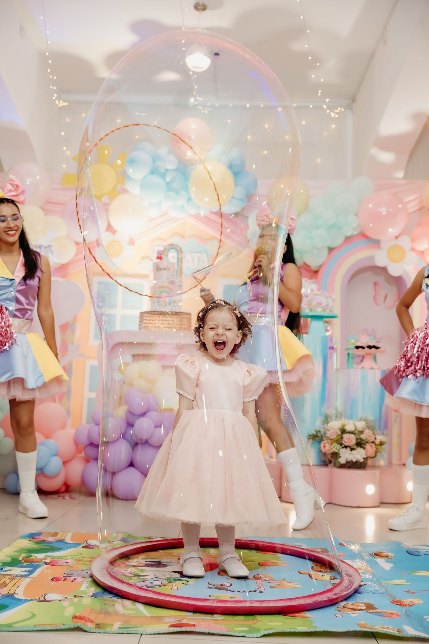 Toddler in white dress jumps joyfully on colorful play mat surrounded by pastel balloons and party decorations.
