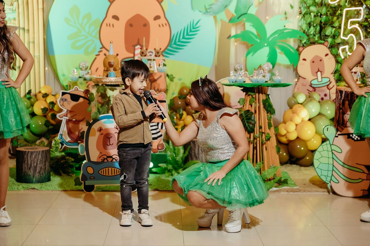 Children celebrate at a dinosaur-themed birthday party with tropical decorations, green balloons, and palm leaf decor.