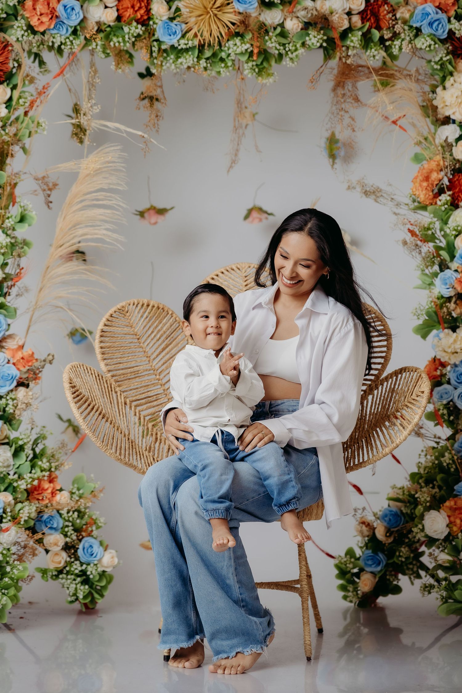 A parent and child sit together in a rattan peacock chair surrounded by a floral arch with roses and pampas grass.