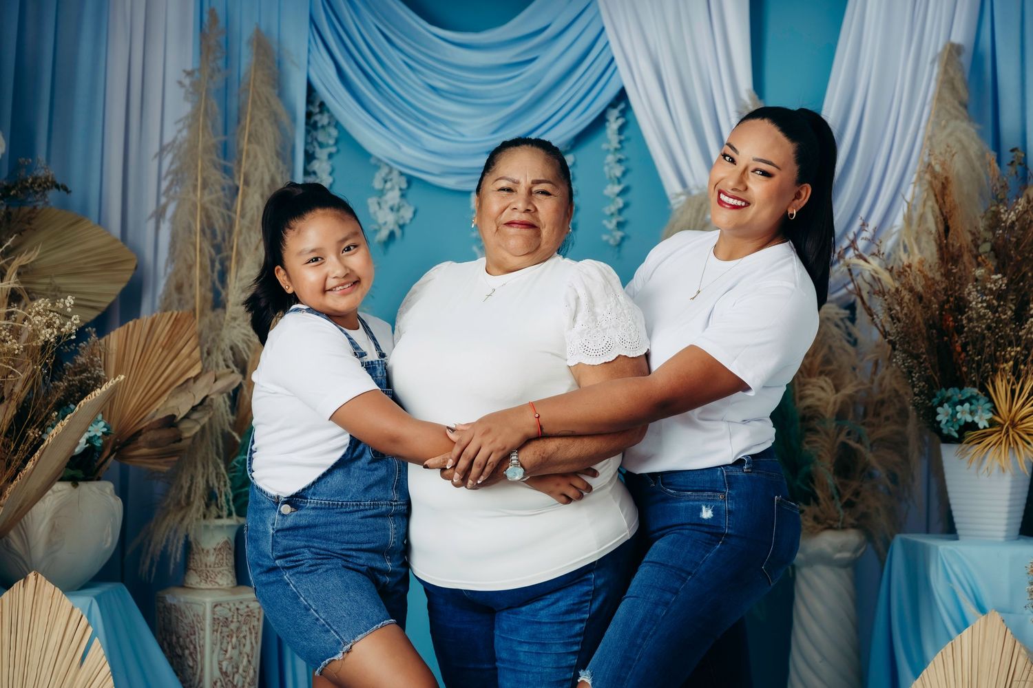 Three women in white tops and jeans smile together in a blue-themed photo studio setting.