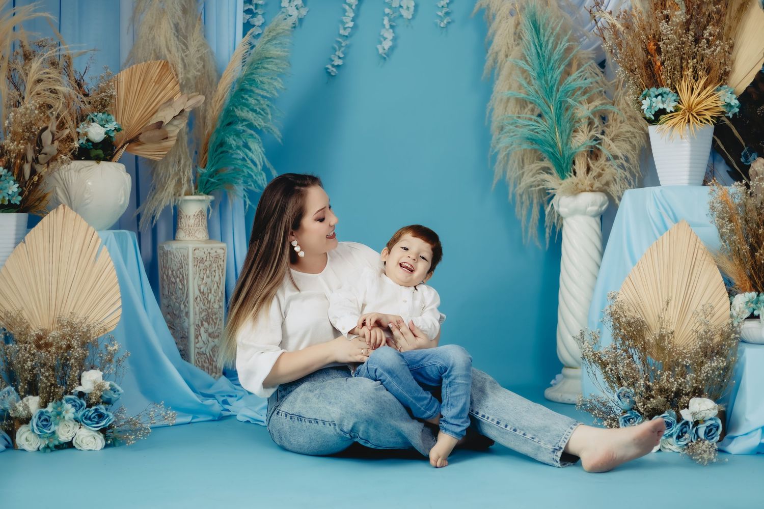 A young mother sits on a blue floor holding her laughing toddler son, surrounded by dried floral arrangements.