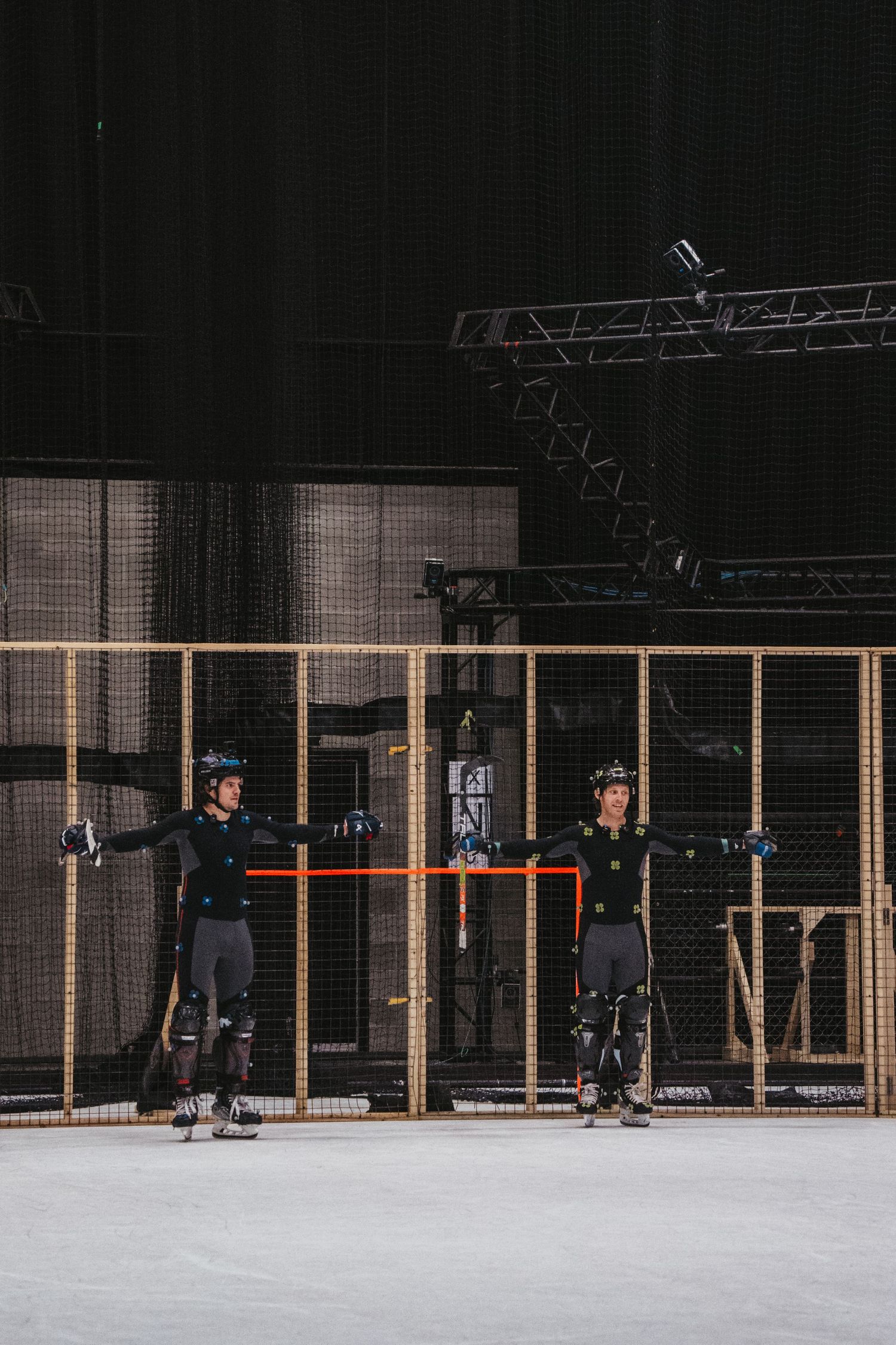 Two performers in motion capture suits stand in front of a wooden set piece on a white floor.