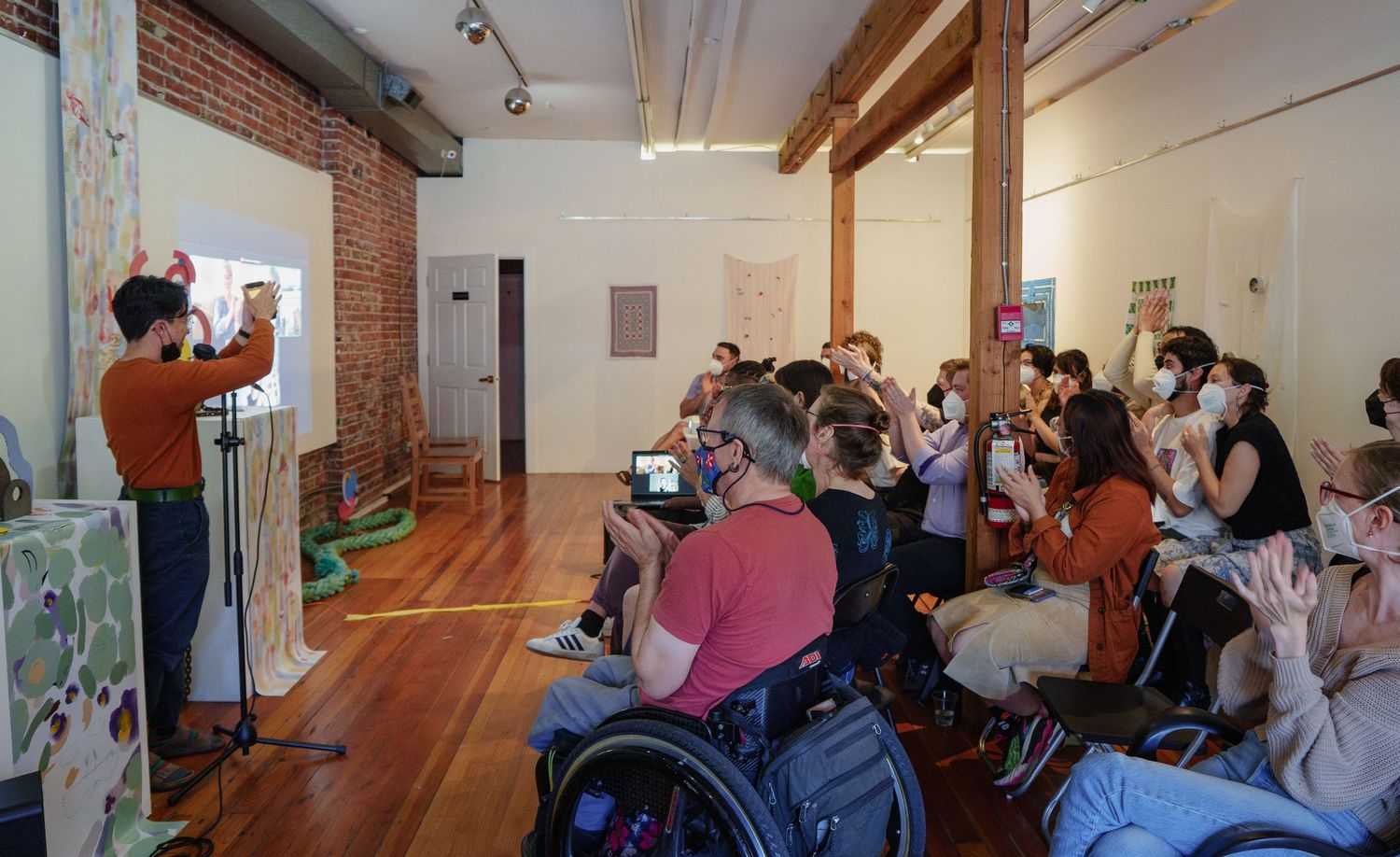 Speaker presents to masked audience in well-lit room with hardwood floors.