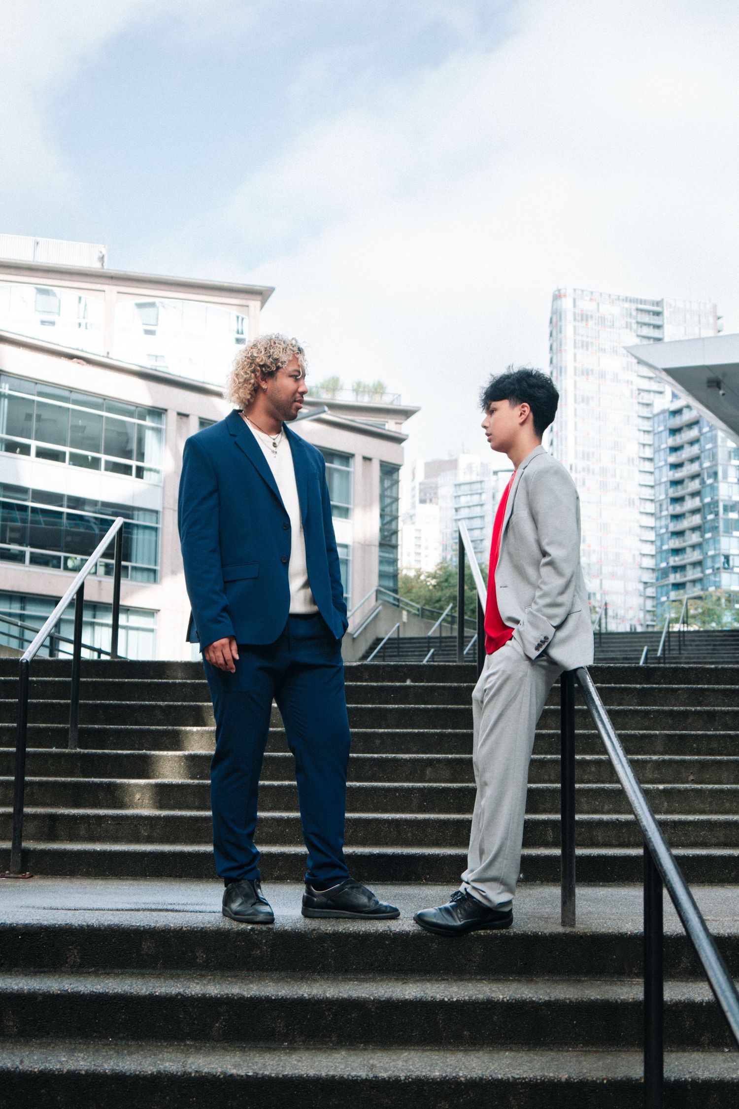 Two models in contrasting suits standing on concrete steps against urban backdrop.