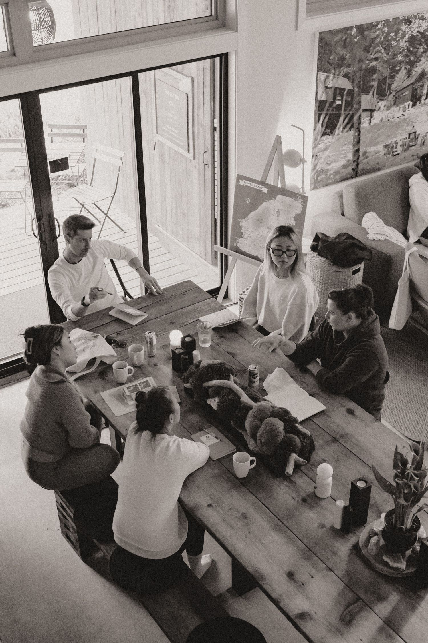 Group gathering around wooden table in bright interior space with large windows.