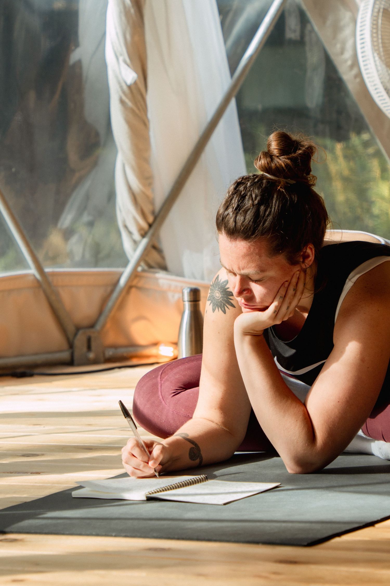 A student writes in journal while lying on yoga mat in sunlit dome studio.