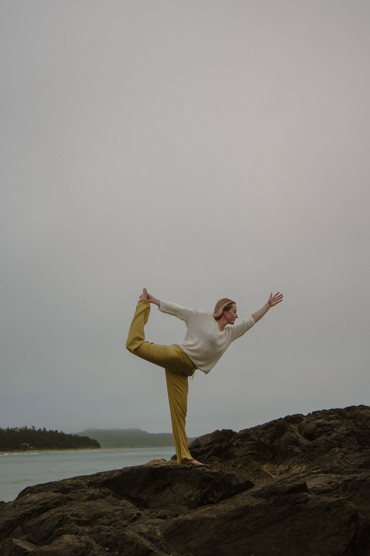 Person performing yoga dancer pose on coastal rocks with ocean backdrop.