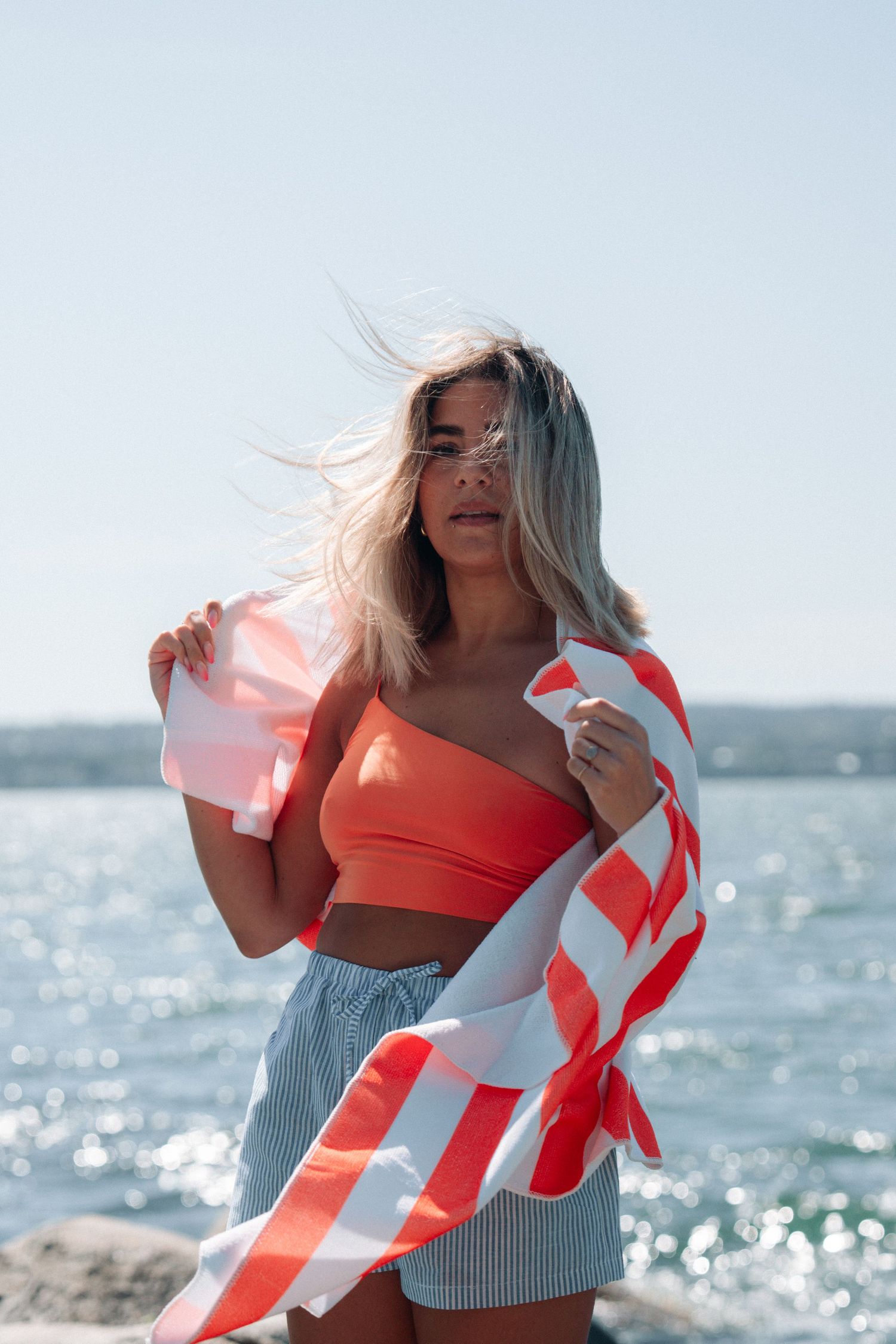 Woman in orange crop top holding striped beach towel by sparkling ocean.