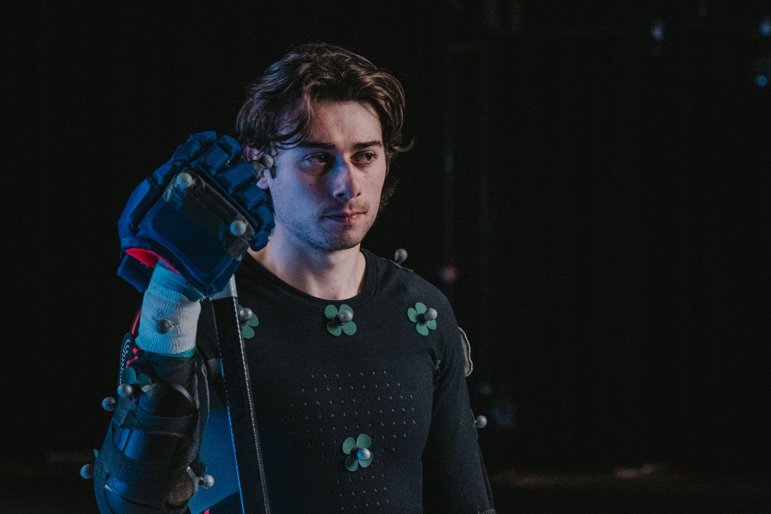 Side view of performer in motion capture gear against dark background with tracking dots.