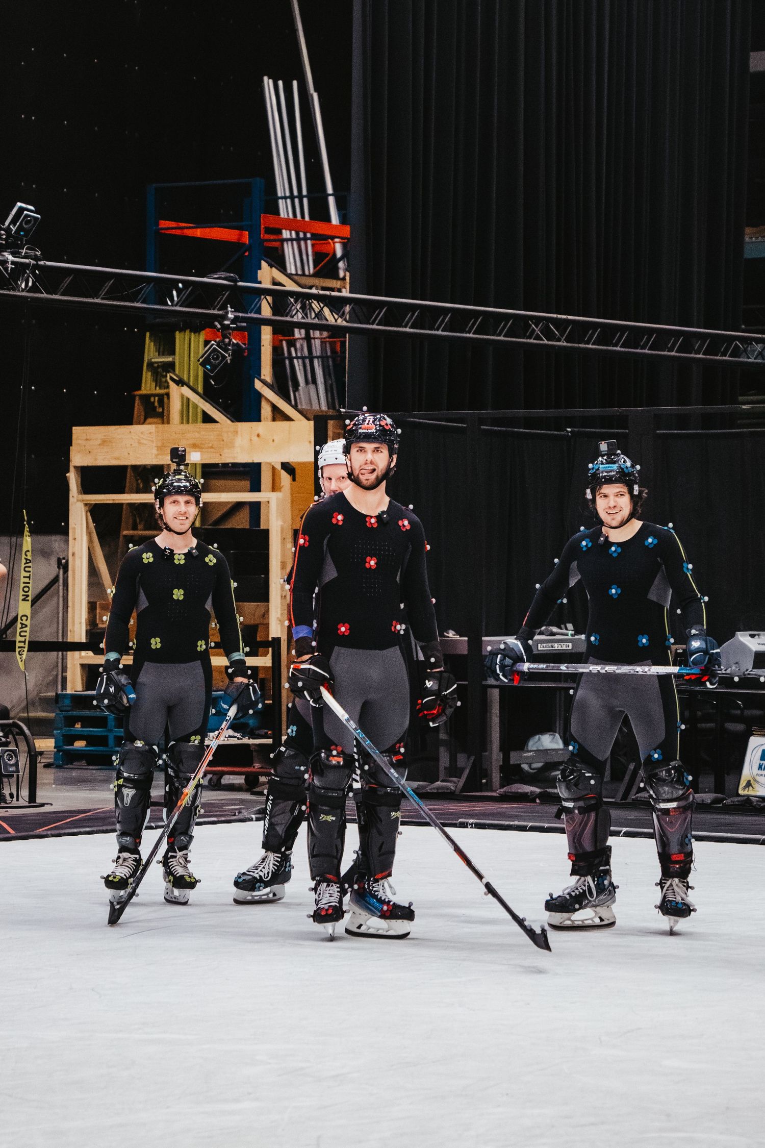 Three performers wearing motion capture suits and holding hockey sticks pose on a white studio floor.