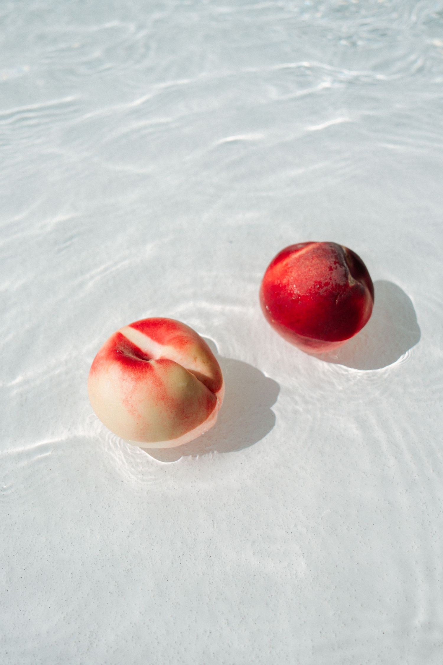 Two fresh peaches casting shadows on white textured surface in bright natural lighting.