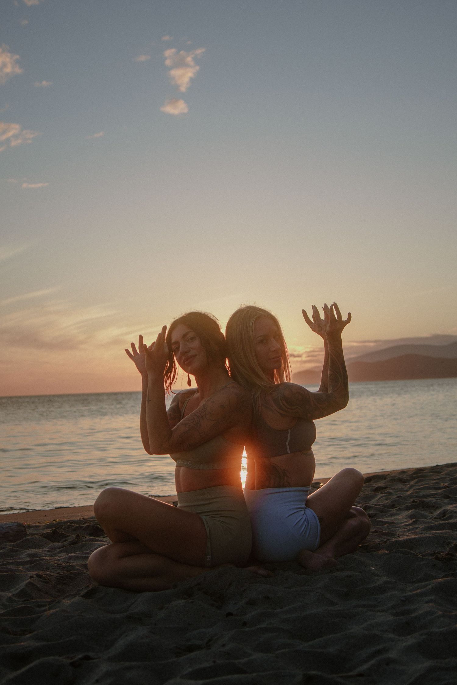 Two figures sitting back-to-back on beach with hands raised playfully at sunset.
