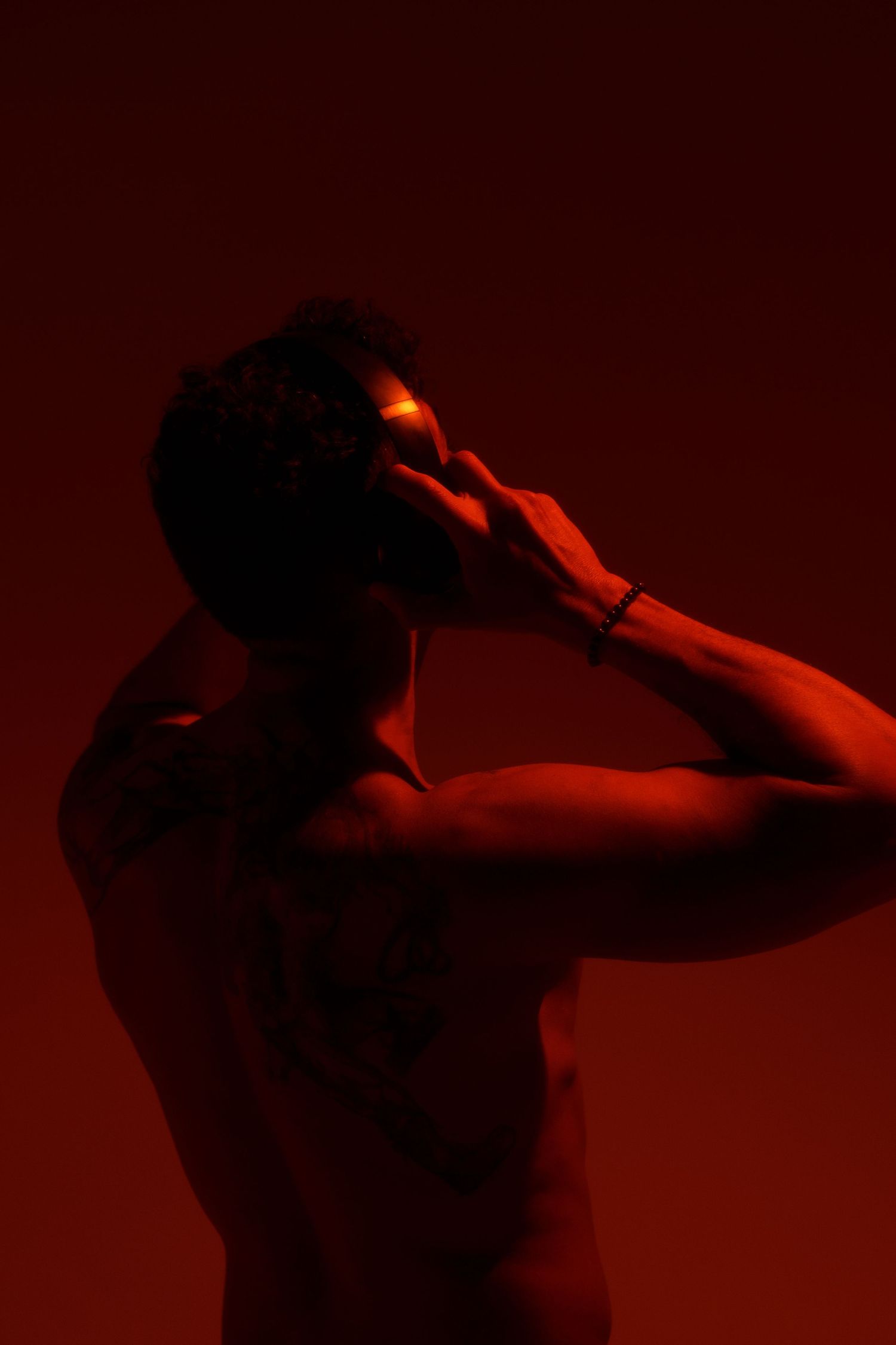 Artistic silhouette of shirtless person lit in dark red lighting with hands raised to head in dramatic pose.