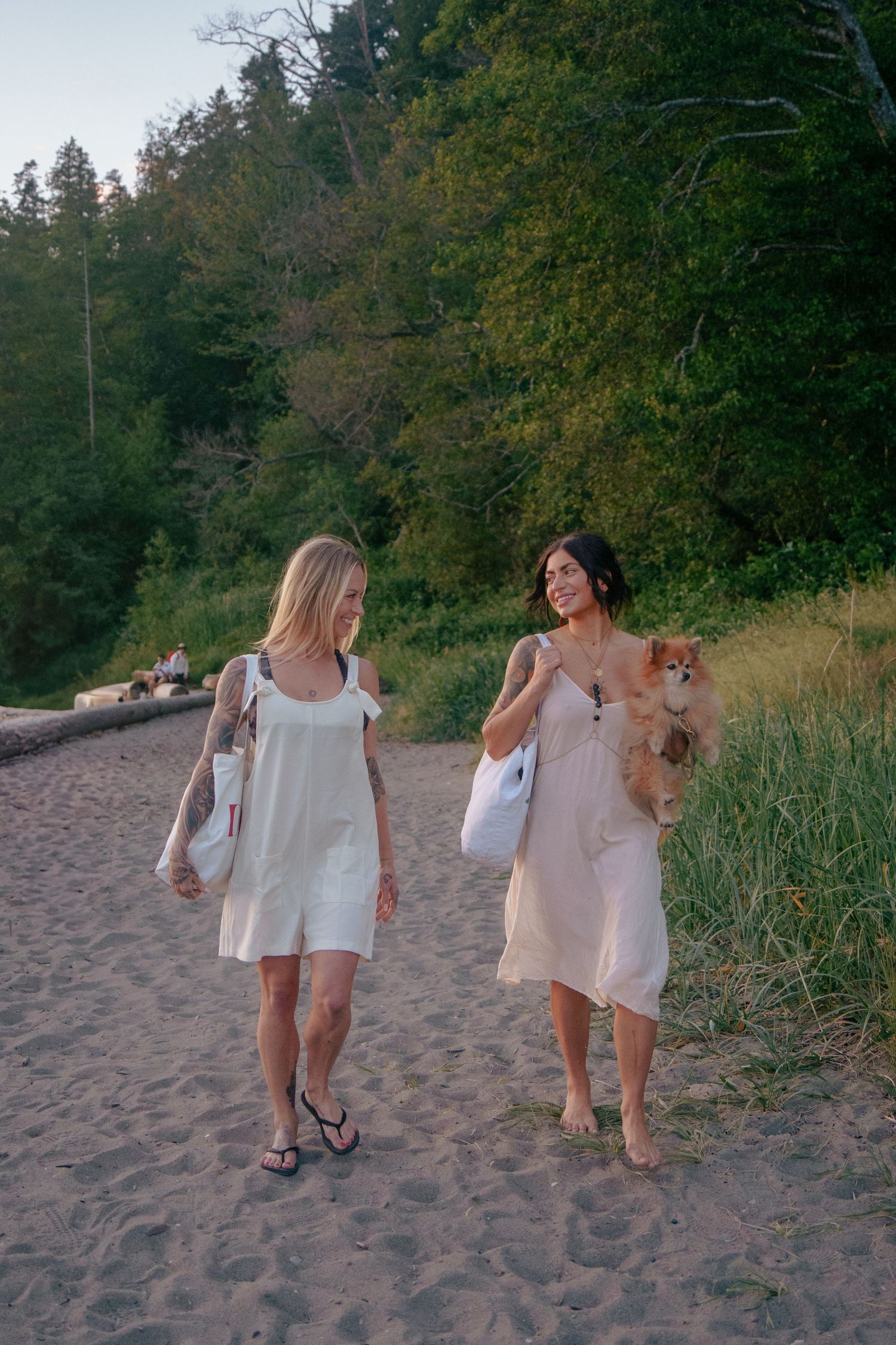 Two friends walking along a sandy beach path carrying white bags near sunset.