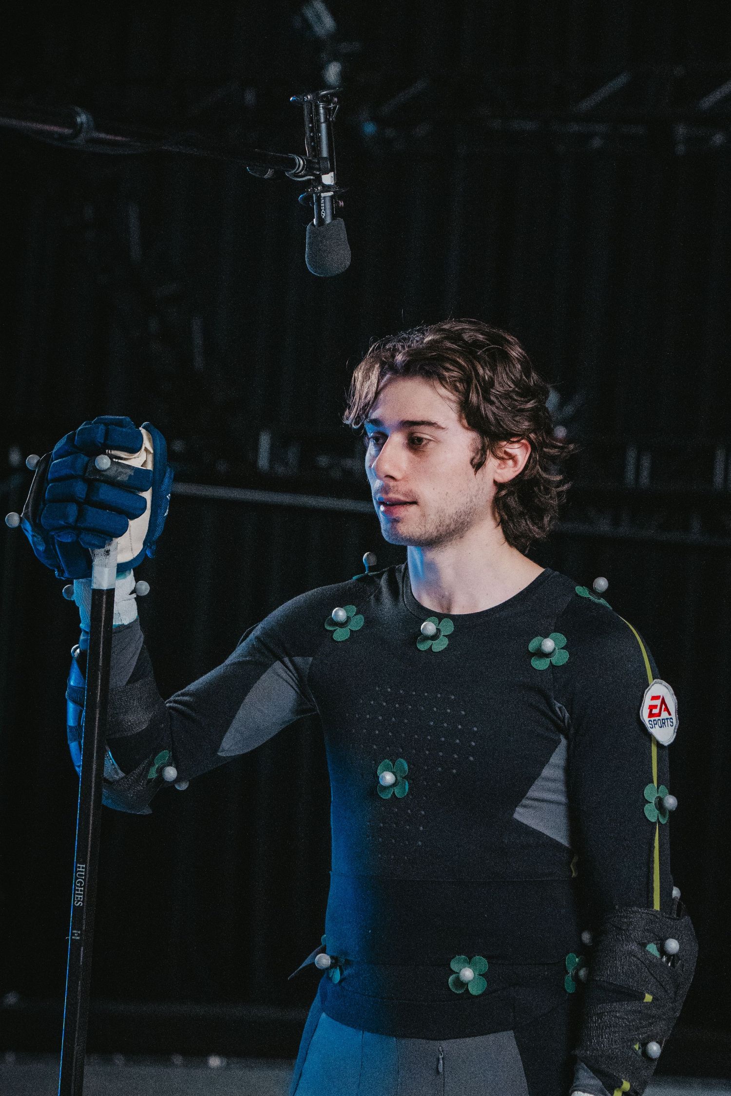 Performer in black suit with green markers stands near microphone during motion capture session.