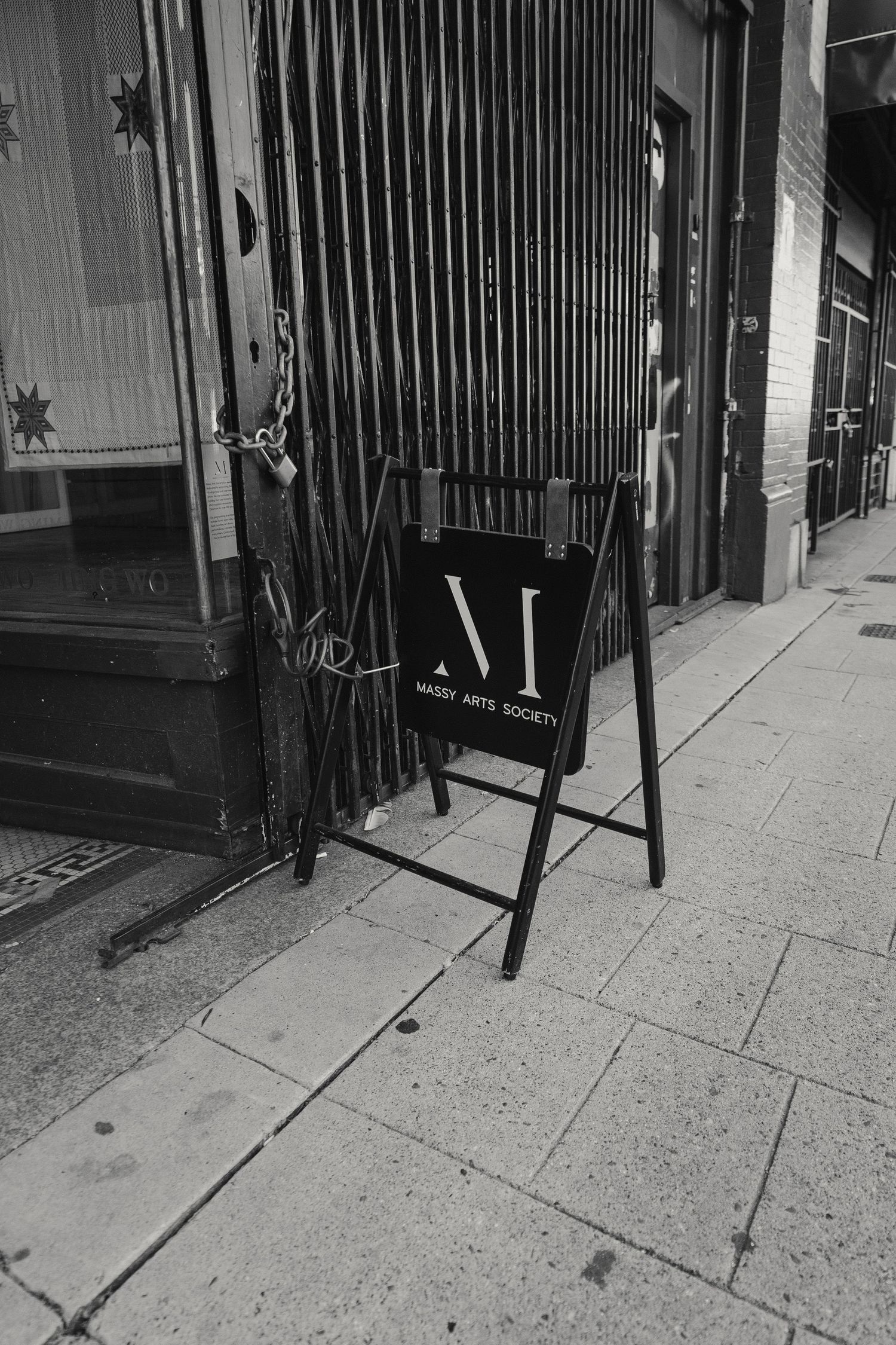 A black and white photo of a metal gate with a signboard on the sidewalk.