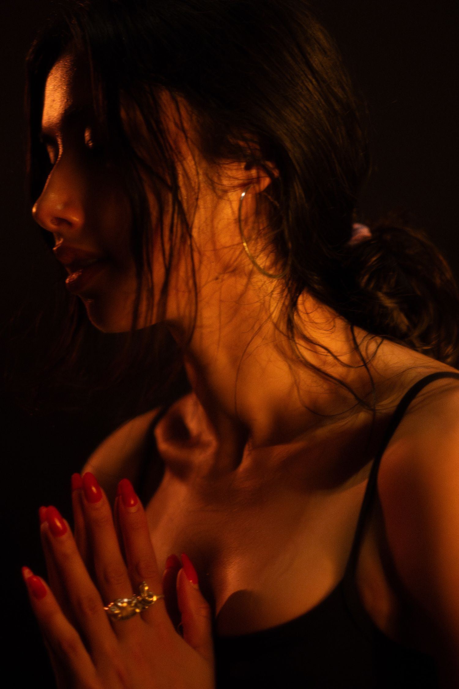 Dramatic side profile portrait with red nails and jewelry illuminated by warm ambient lighting.