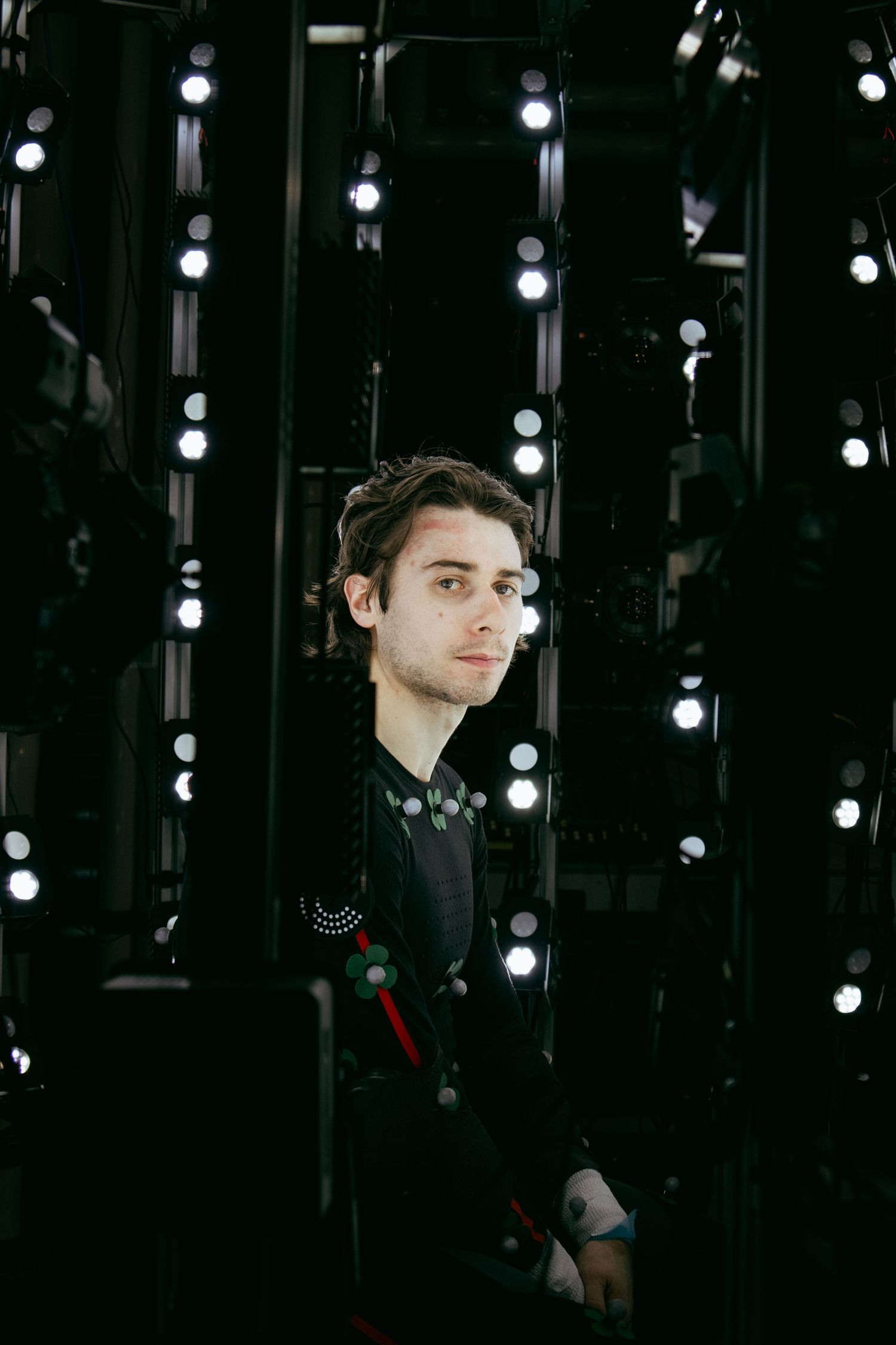Motion capture performer in black suit stands among vertical rows of studio lighting.