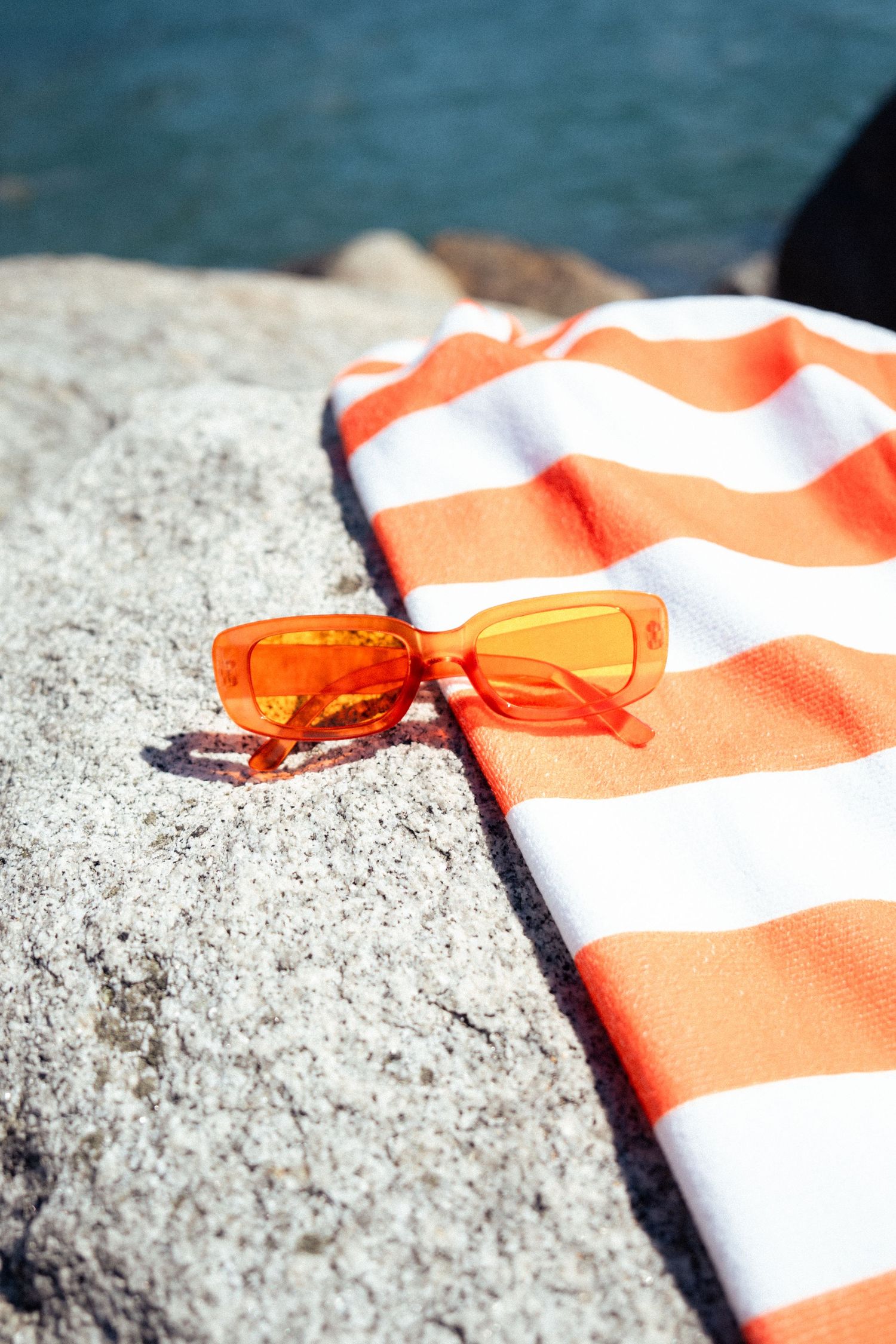 Orange sunglasses on striped beach towel against rocky coastal background.