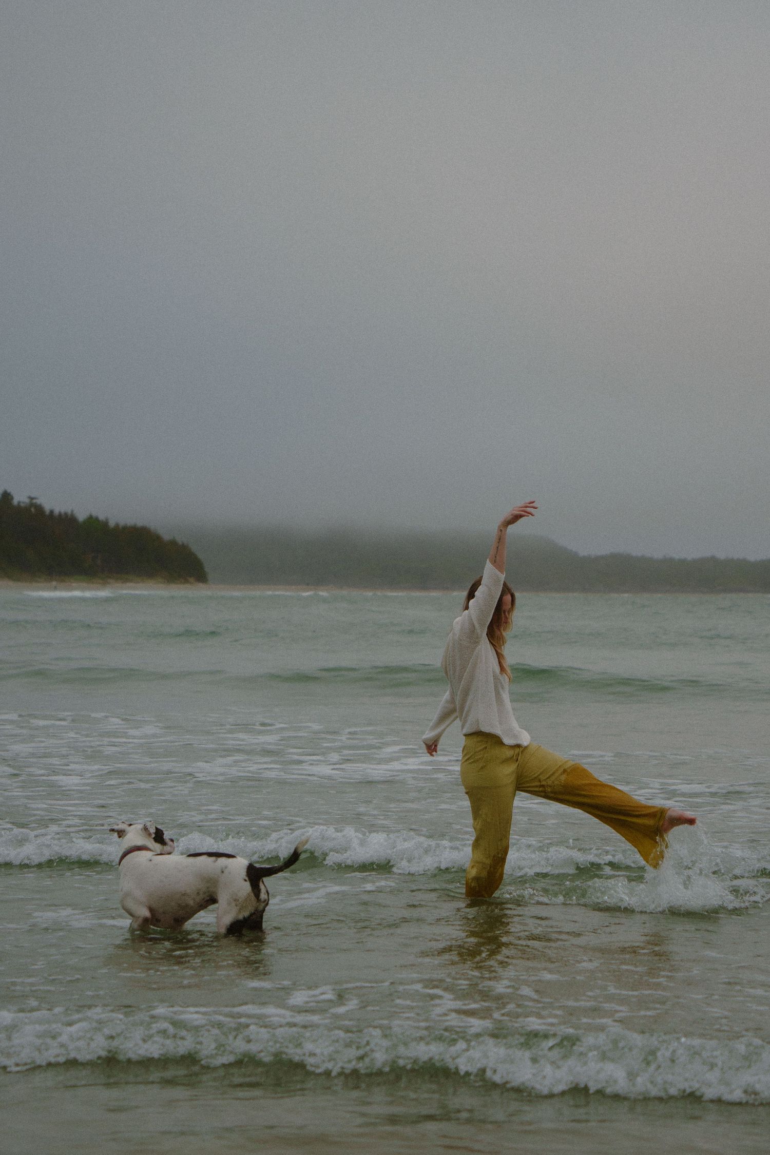 Person in yellow pants dancing in ocean waves with a dog playing nearby.