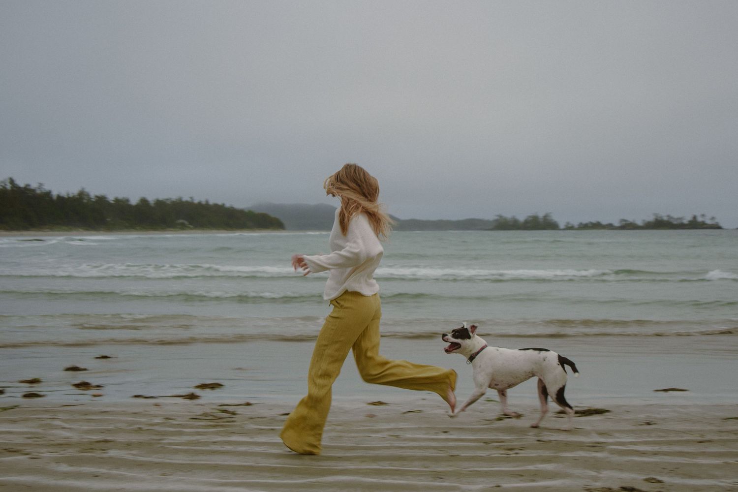 Person in yellow pants running joyfully along a beach with their dog on an overcast day.