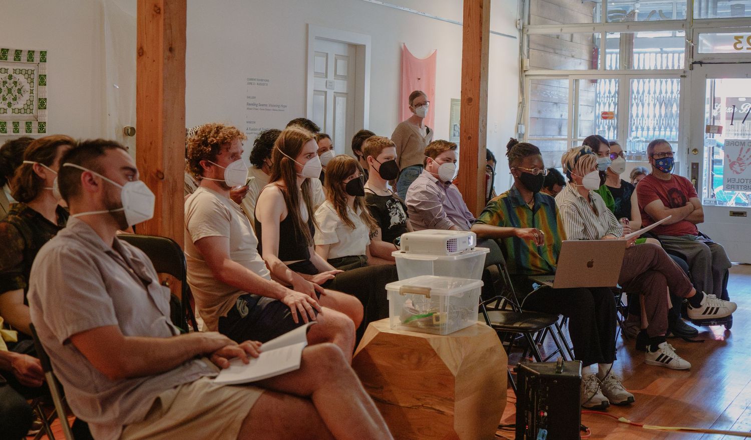 A casual audience sits in rows during an indoor event with exposed wooden beams and brick walls.