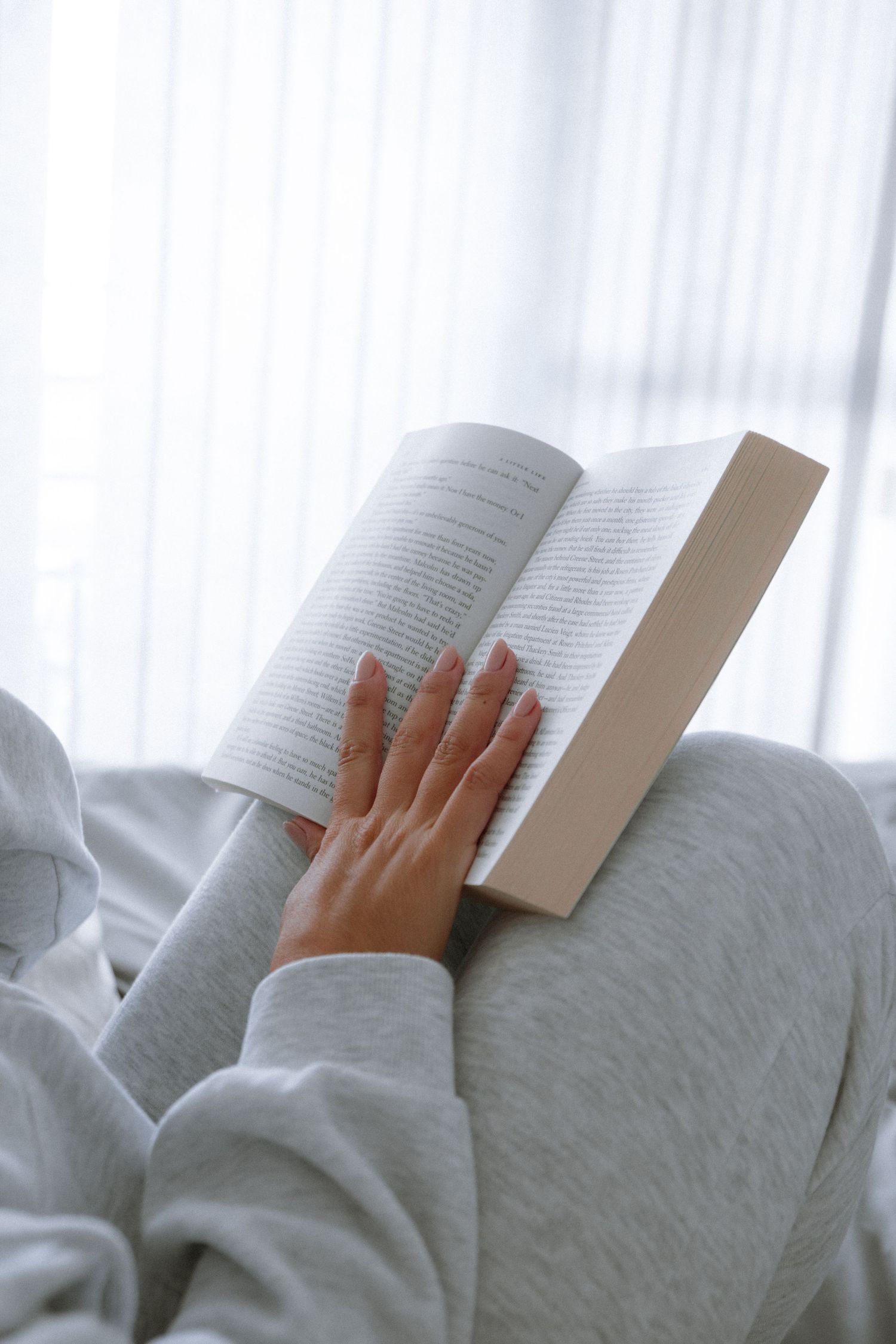 Hands holding an open book against white bedding in natural light.