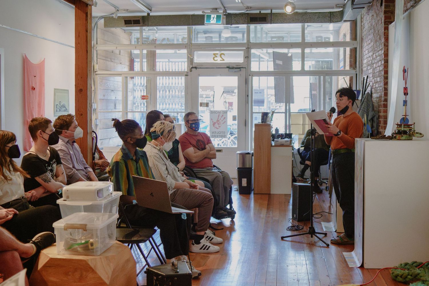 An audience watches attentively during an indoor performance in a bright room with wooden floors.
